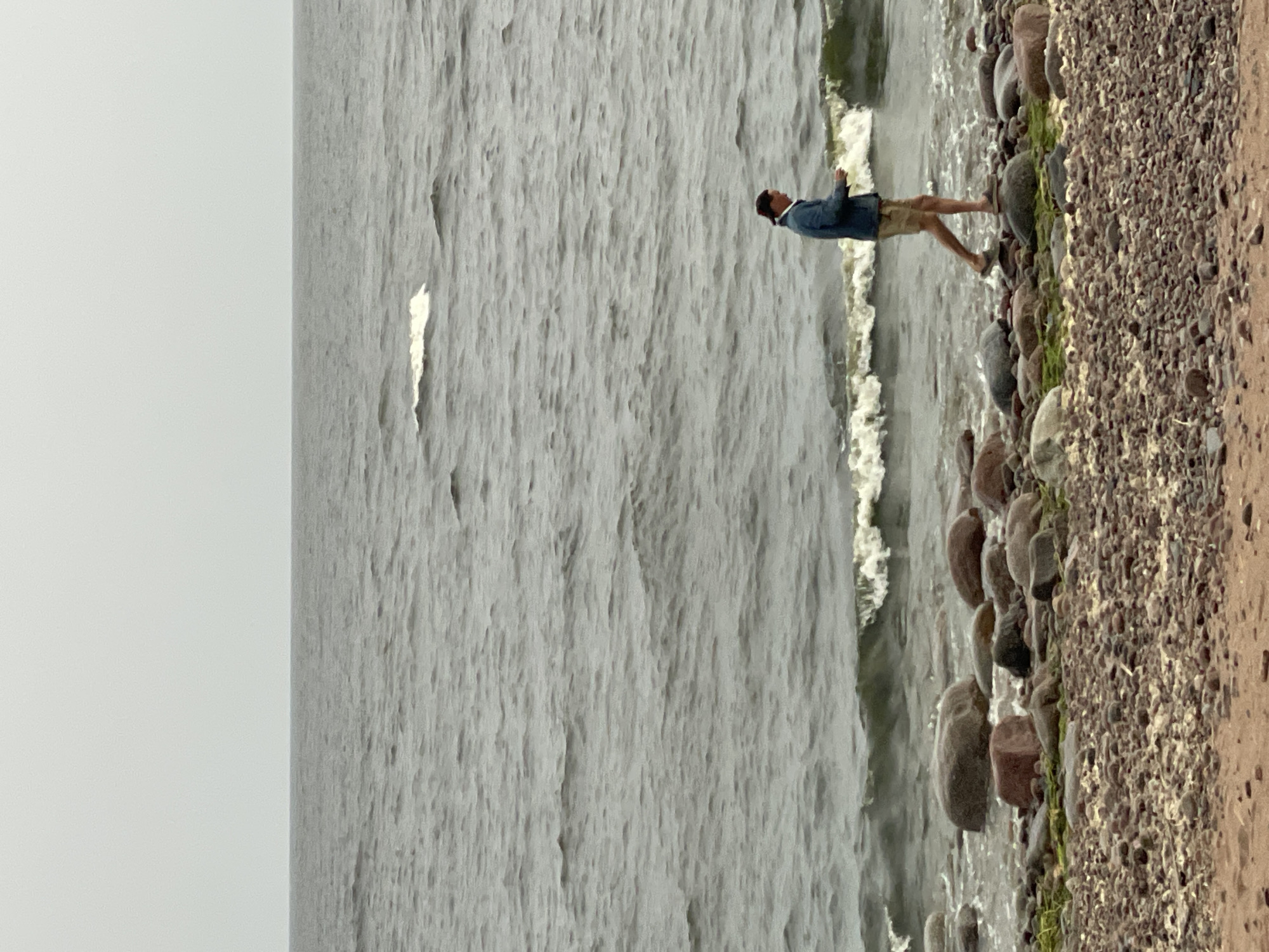 A man walks along the rocky shore of the Gulf of Finland in Luzhki, Russia, on a cloudy day. The man, wearing a blue jacket and light-colored shorts, is captured in profile, his gaze directed downwards. He is walking amidst a scattering of rounded rocks of various sizes, some partially submerged in the water, with a foamy wave breaking behind him. The sea stretches out to the horizon under a pale, overcast sky. The foreground is a mix of sand and pebbles.