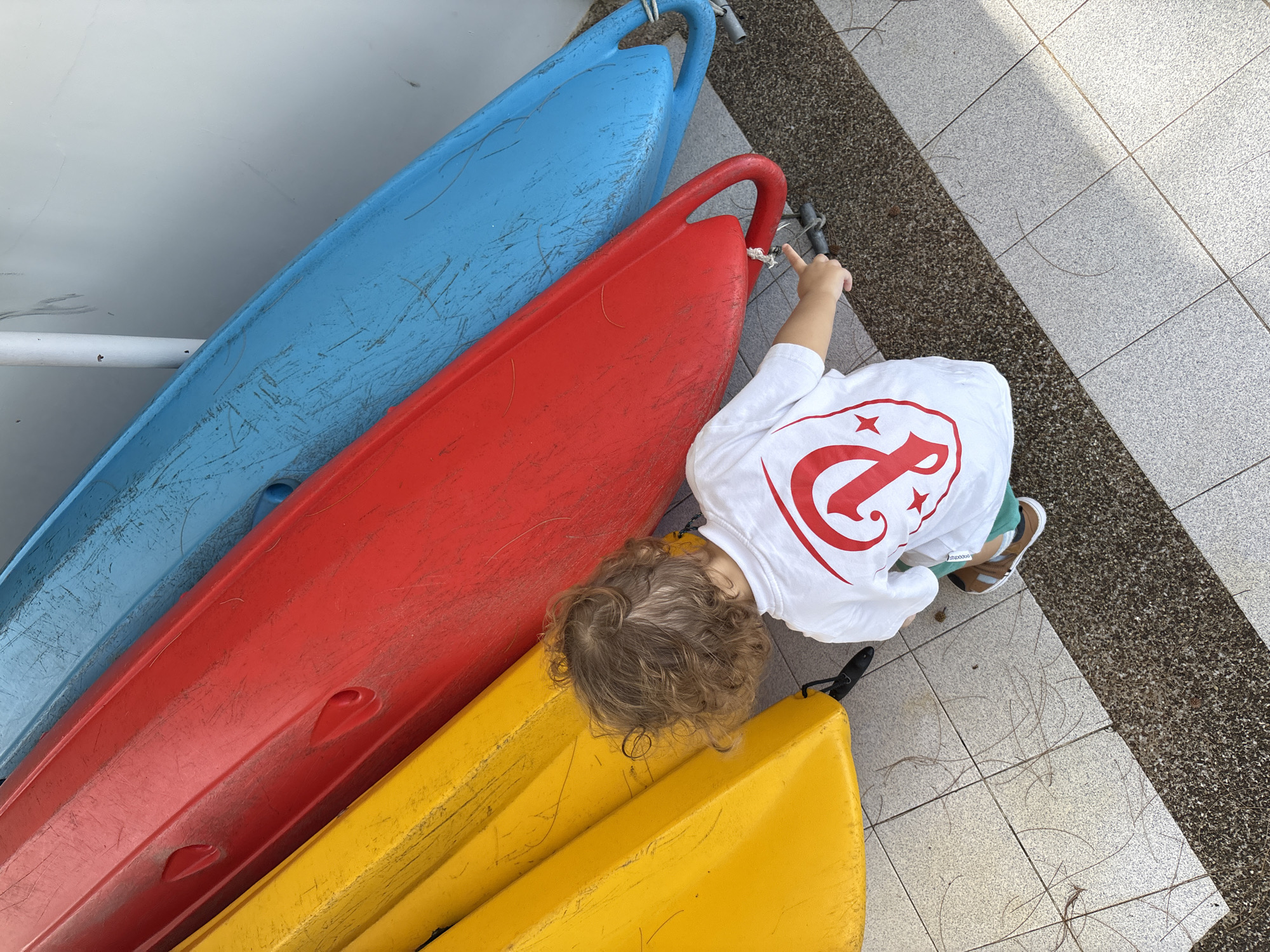 In Choeng Thale, Thailand, a young child with curly brown hair kneels on a tiled outdoor surface, facing away from the camera. The child is wearing a white t-shirt with a red graphic design and green shorts, with brown sandals on their feet. They are pointing a finger towards a red kayak, which is leaning against a blue kayak. Another yellow kayak is positioned in front of the red one. The kayaks are situated next to a light gray wall. The ground is a combination of light-colored tiles and a darker, speckled material. Pine needles are scattered across the tiled surface.