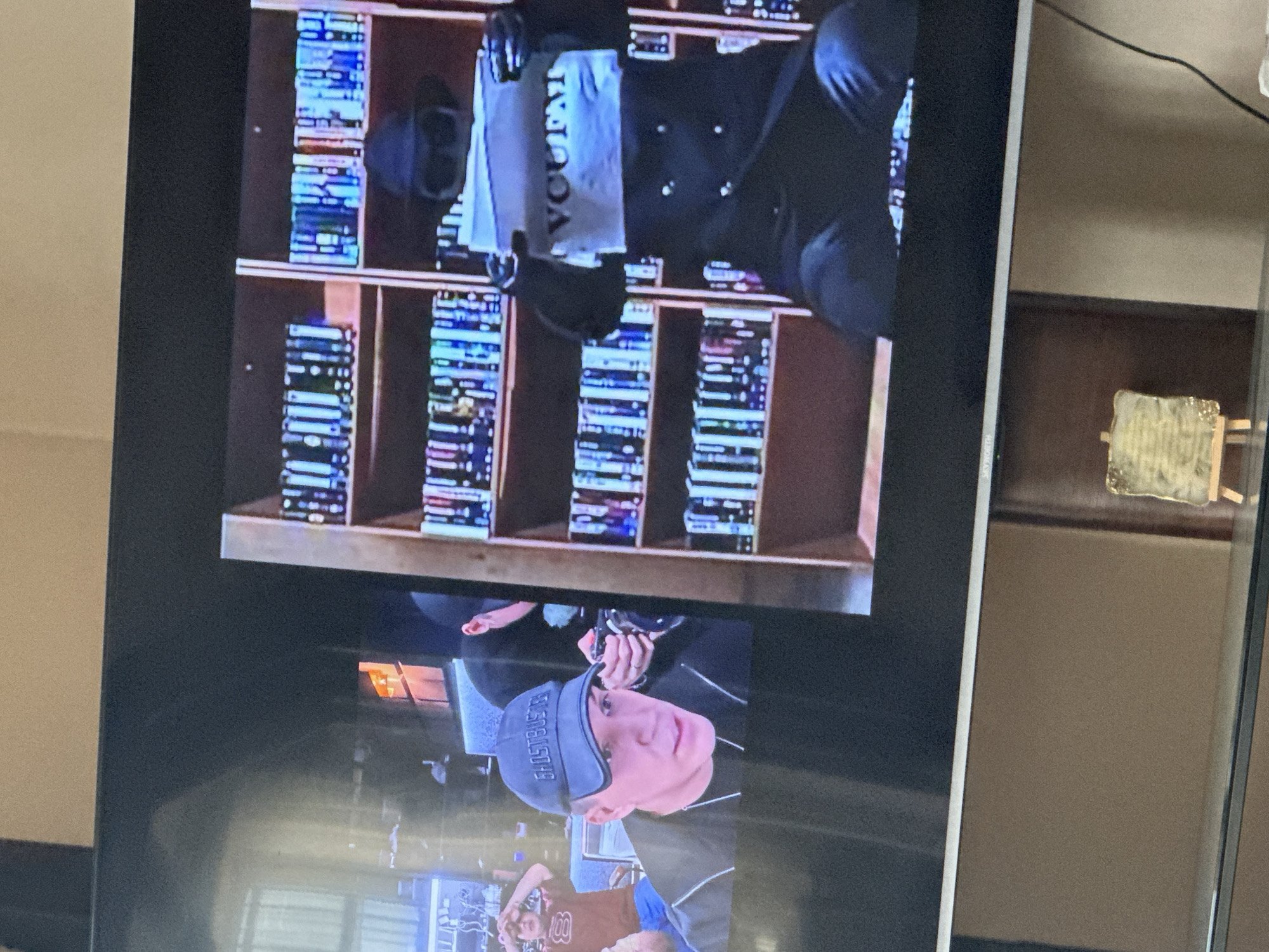 The image shows a television screen displaying a scene. On the screen, there's a shelf filled with what appears to be video tapes or DVDs, arranged neatly. In front of the shelf, a person is seen wearing a black outfit and holding what looks like a white t-shirt with black text. In the lower part of the screen, there's another person wearing a "GHOSTBUSTERS" cap and a jersey.