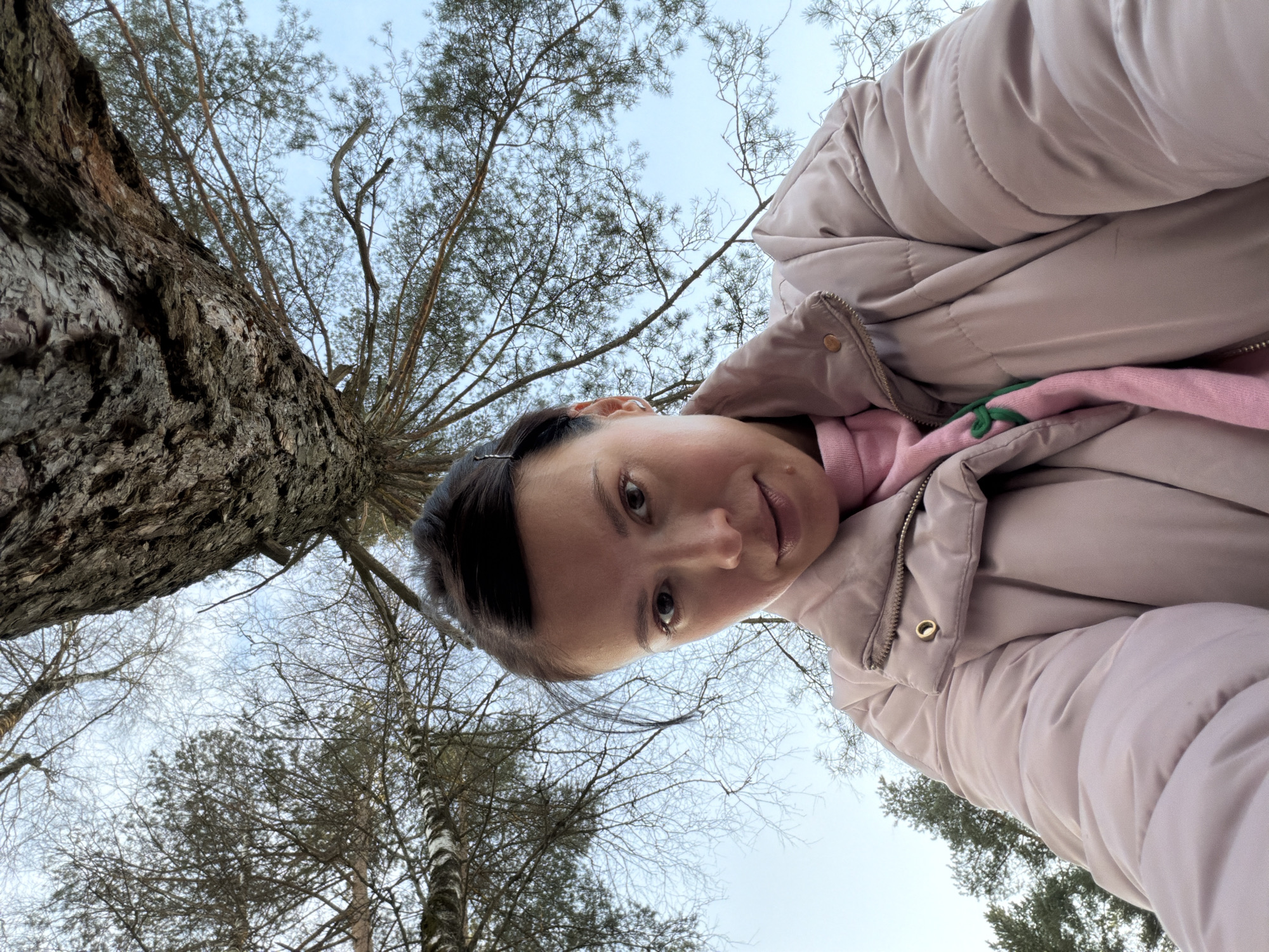 An adult female is depicted in an outdoor setting within a wooded area in Khimki, Russia. The image is captured from a low, upward-facing angle, placing her in the foreground looking directly at the camera with a neutral expression. She wears a light pink puffer jacket over a pink sweater, which exhibits a small green knot or detail near the collar. Her dark hair is pulled back, secured by a small hair clip on the left side of her head, and a metallic earring is visible on her left earlobe. The background consists of a partially overcast pale blue sky and a dense canopy of trees. Prominent features include a large tree trunk with dark, rugged bark in the upper right, multiple deciduous trees with bare branches, and evergreen conifers with needles, suggesting a mixed forest environment in cooler weather.