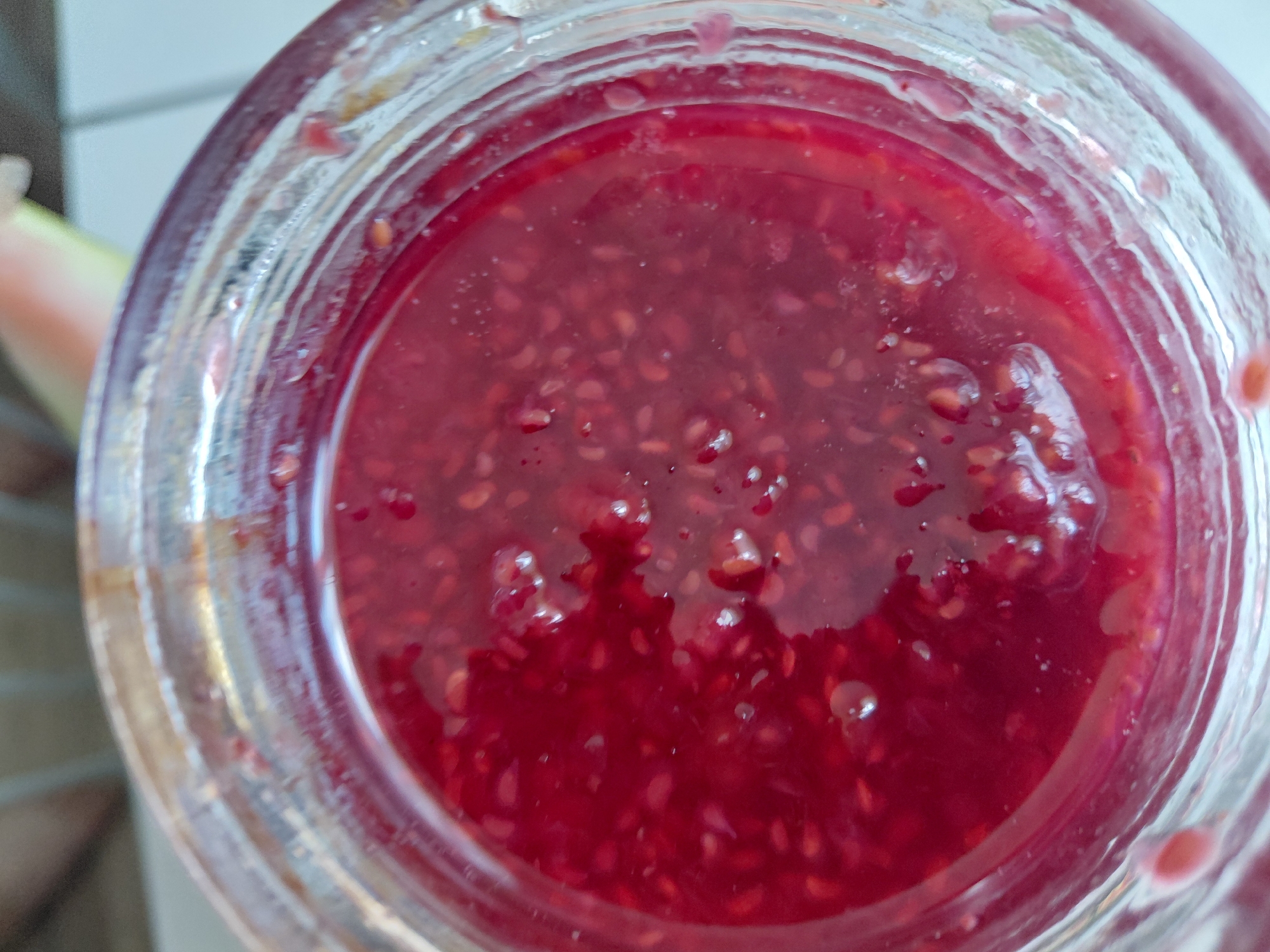 A close-up, overhead shot shows a glass jar filled with raspberry jam. The jam is a deep red color with visible seeds scattered throughout. The glass jar has ridged detailing around the rim. The background is blurred and out of focus, with hints of light colors and textures that do not provide any specific environmental context. The scene appears to be indoors. There are no people or actions depicted.