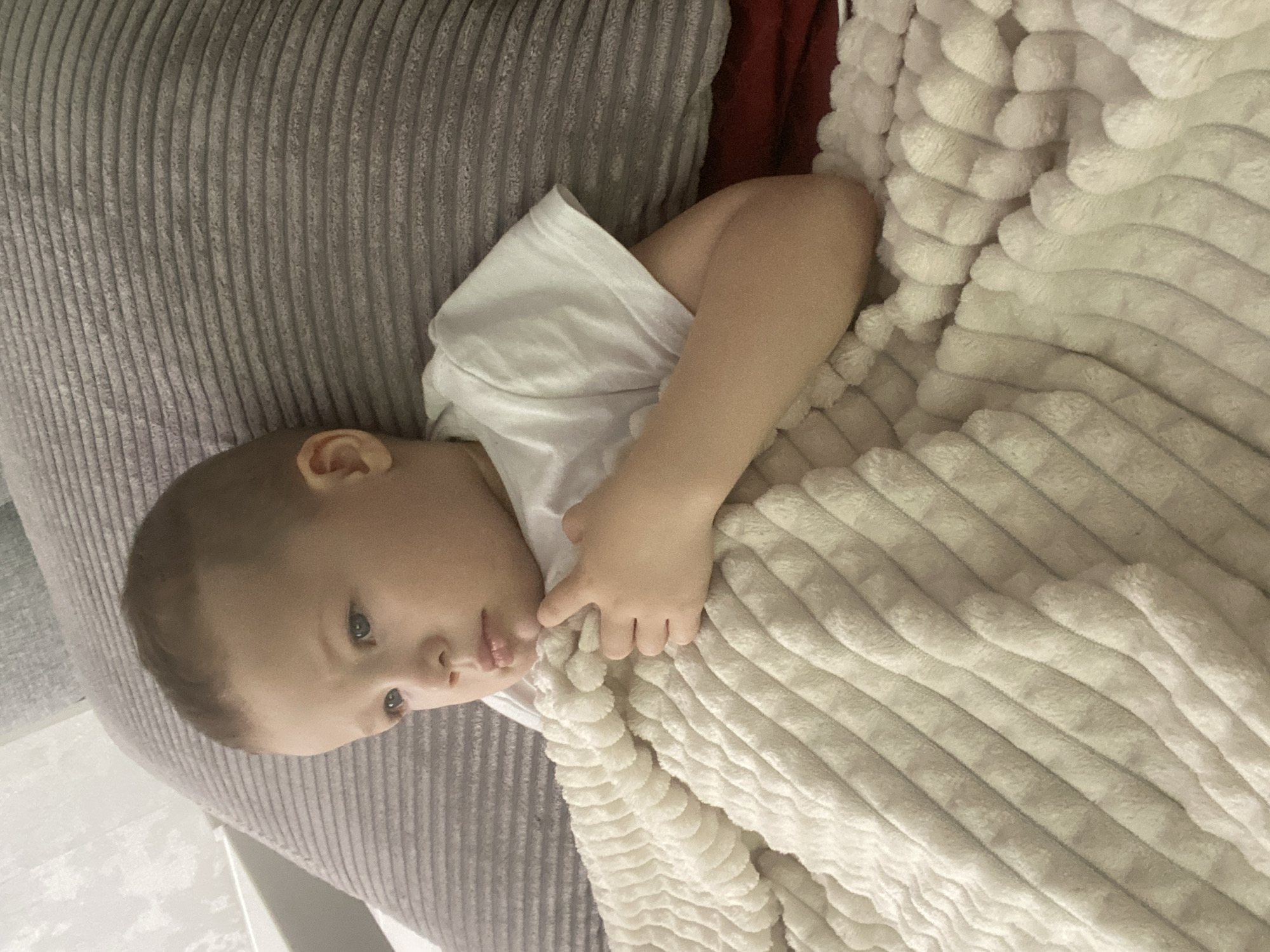 A young child, likely between 1-3 years old, is lying on a gray corduroy sofa or cushion and partly covered with a soft, light-colored blanket with a horizontally ribbed texture. The child is wearing a white t-shirt. The child's face is visible, and they appear to be resting. A small portion of a red object is visible behind the child and to the left of the blanket. There's no indication of the image being taken in Sochi, Russia.