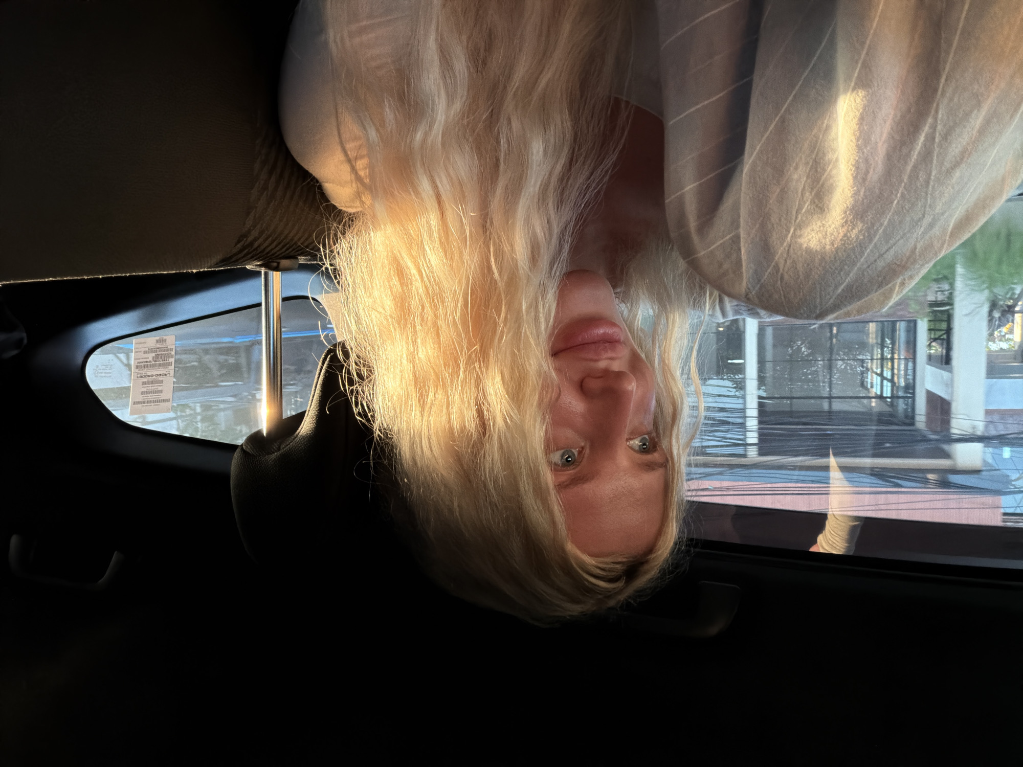A young blonde woman is photographed from the chest up, seated inside a vehicle in North Kuta, Indonesia. The woman is looking to her right, with sunlight illuminating her wavy blonde hair and the right side of her face. She is wearing a white top, and a cream-colored garment with thin white stripes is draped over her left shoulder and lap. The interior of the vehicle is dark, with the dark headrest of a seat visible behind her. Through the window, a blurred cityscape is visible, characterized by buildings and overhead wires, with sunlight reflecting off some surfaces. A white sticker with barcodes and text is affixed to the window. The overall lighting suggests it is late afternoon or early evening.