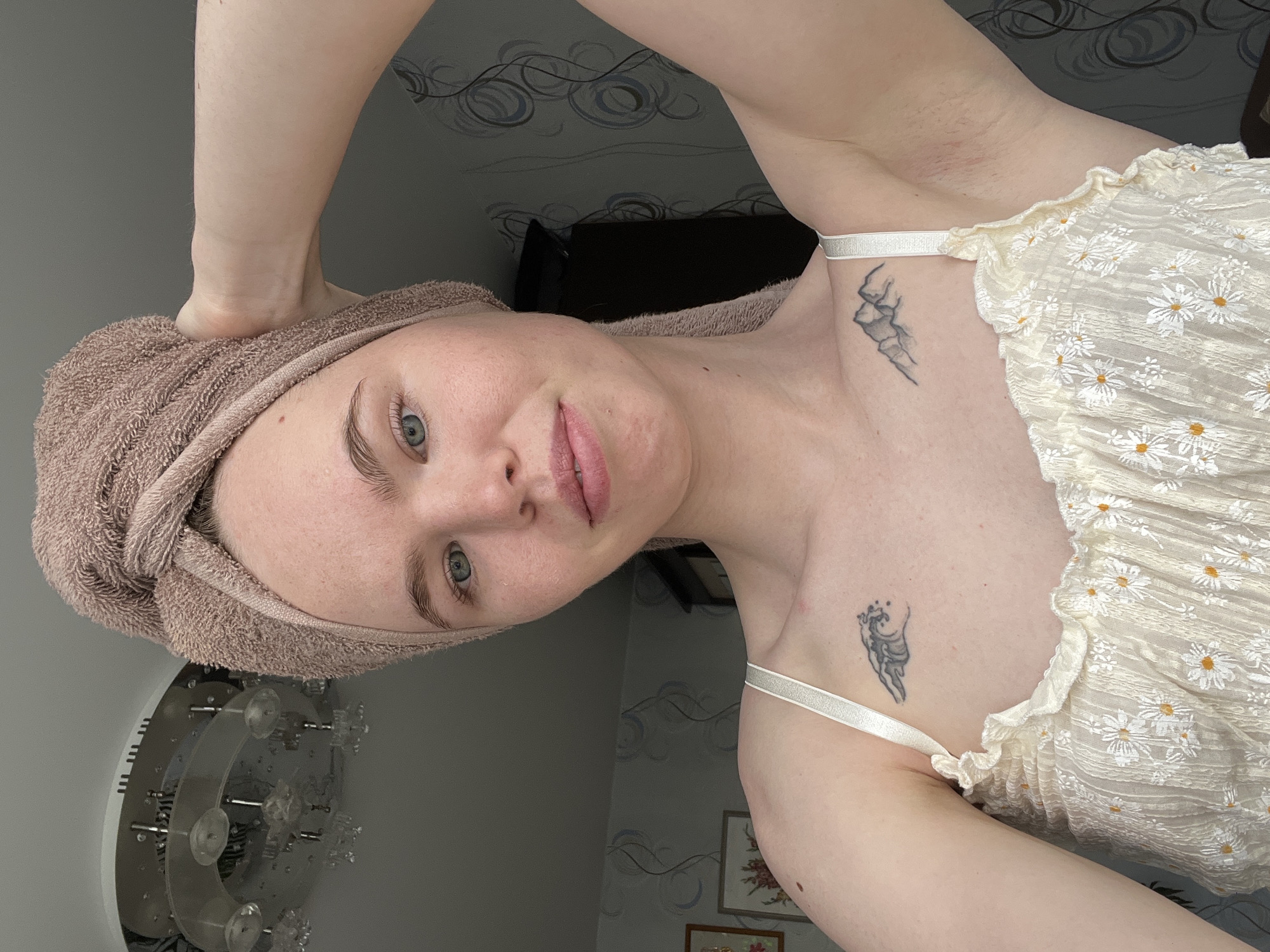 A selfie of a woman in a room in Chaikovsky, Russia. She has fair skin, blue eyes, and light eyebrows, with a brown towel wrapped around her head. She is wearing a white top with daisy patterns and thin straps. Tattoos are visible on her upper chest/shoulder area. The background shows a wall with patterned wallpaper, a partial view of a dark object, and a circular, decorative object on the wall.