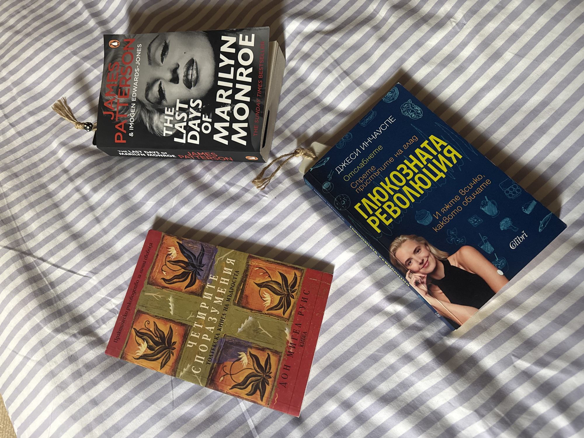 The image shows three books placed on a striped bedsheet. The book on the top left is "The Last Days of Marilyn Monroe" by James Patterson and Imogen Edwards-Jones. The book on the top right has a blue cover and is in Bulgarian, with a woman on the cover, and the title appears to be "Глюкозната Революция". The book on the bottom is also in Bulgarian with an ornate cover, the title appears to be "Четирите споразумения". All three books are laying flat and appear to have bookmarks inside. The background is a striped bedsheet.