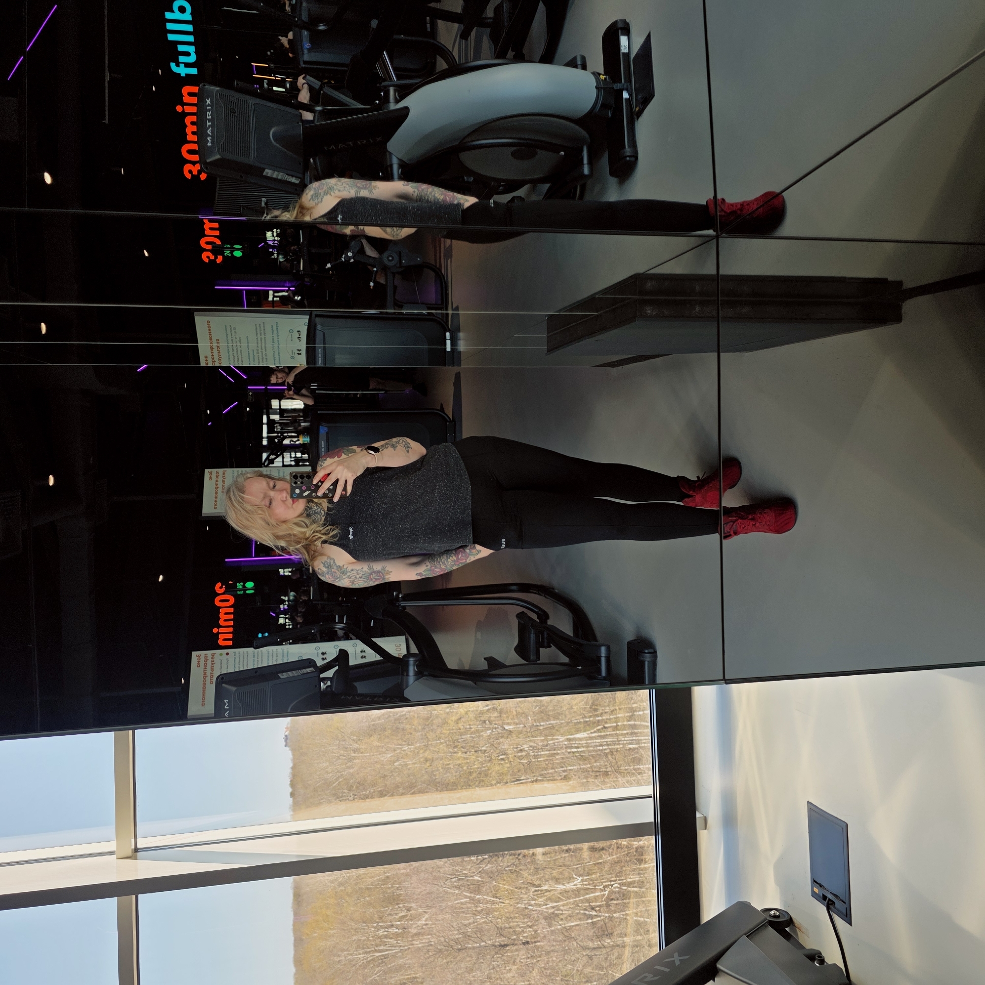 The image captures a full-length mirror selfie of a woman in a gym, set in Saint Petersburg, Russia. The woman, with blonde hair and visible arm tattoos, is wearing a dark grey tank top, black leggings with a white stripe, and bright red athletic shoes. She holds a smartphone up to take the photo, reflecting herself and the gym environment. Her expression is calm, with a slight smile.
The setting is a modern fitness center with dark, highly reflective walls, giving the impression of multiple mirrors or glass partitions. Several pieces of gym equipment are visible, including what appear to be stationary bikes or elliptical machines, one bearing the "MATRIX" brand name. Neon purple lighting accents are visible in the background, contrasting with the dark interior.
Below the reflective surface, a large window reveals an outdoor scene. The view is of a wooded area with bare trees, suggesting late autumn, winter, or early spring. The bright sky indicates it is daytime.
Visible text elements include "30min fullb" in bright blue and red, and "30m" (likely referring to workout duration) on signs within the gym. Cyrillic text, some of which is reversed due to reflection, includes "ЗОНА" (ZONE) and "ОДОБРЕННОГО РЕЗУЛЬТАТА" (OF THE APPROVED RESULT), likely part of informational signage. The extensive reflective surfaces create a dynamic and somewhat complex visual composition.