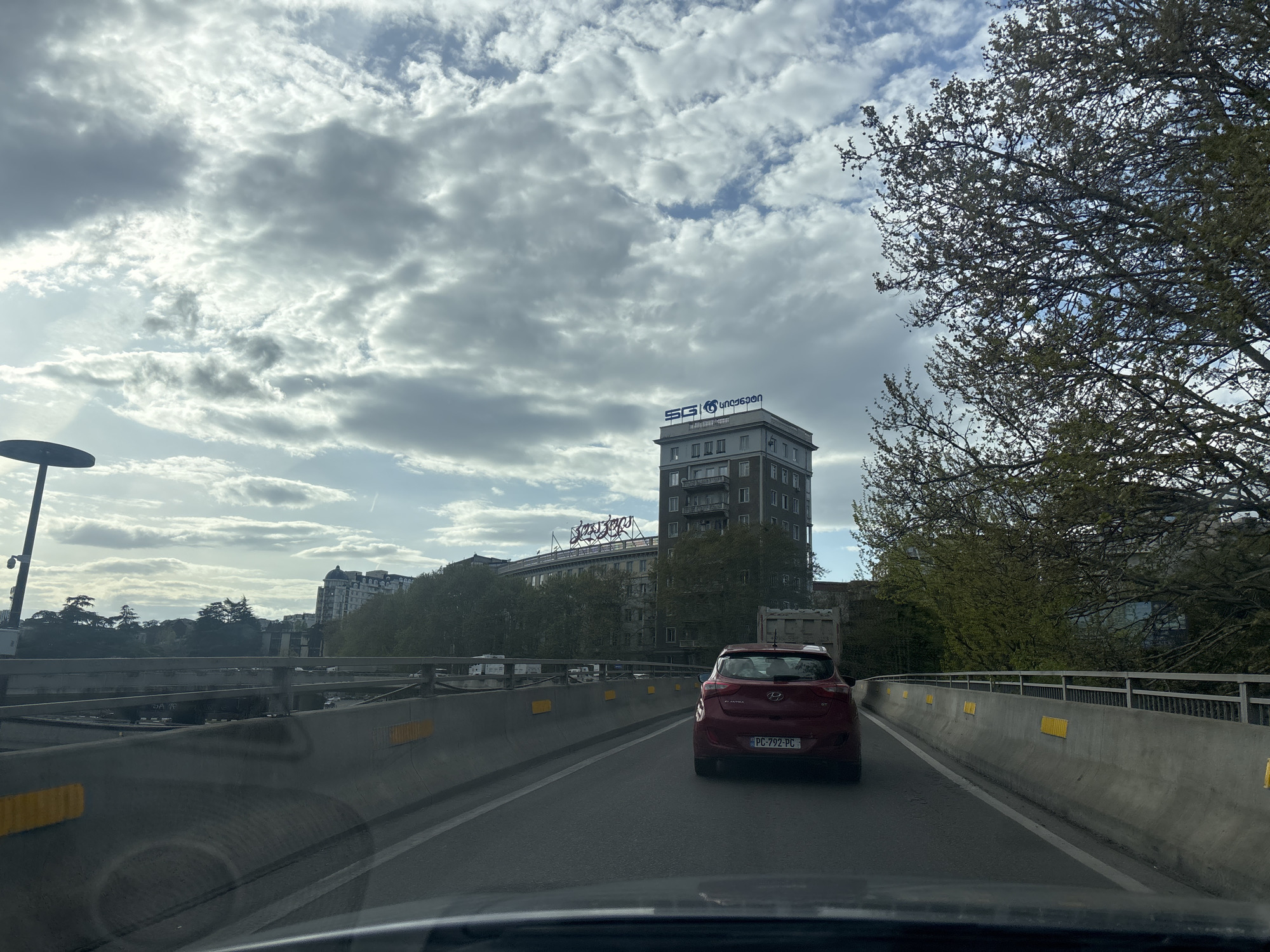 A photograph taken from inside a moving vehicle shows a red Hyundai Elantra GT (license plate PC-792-PC) driving on a highway bridge in Tbilisi, Georgia. The vehicle is on the right-hand side of the road. The road is surrounded by concrete barriers with intermittent yellow reflectors. In the background, there is a tall building with a blue "SG" logo and Georgian text on top. Another building with signage in Georgian is partially visible below the SG building. Trees with budding leaves are visible to the right and left of the buildings. The sky is cloudy with patches of blue. The vehicle's dashboard is visible in the foreground.