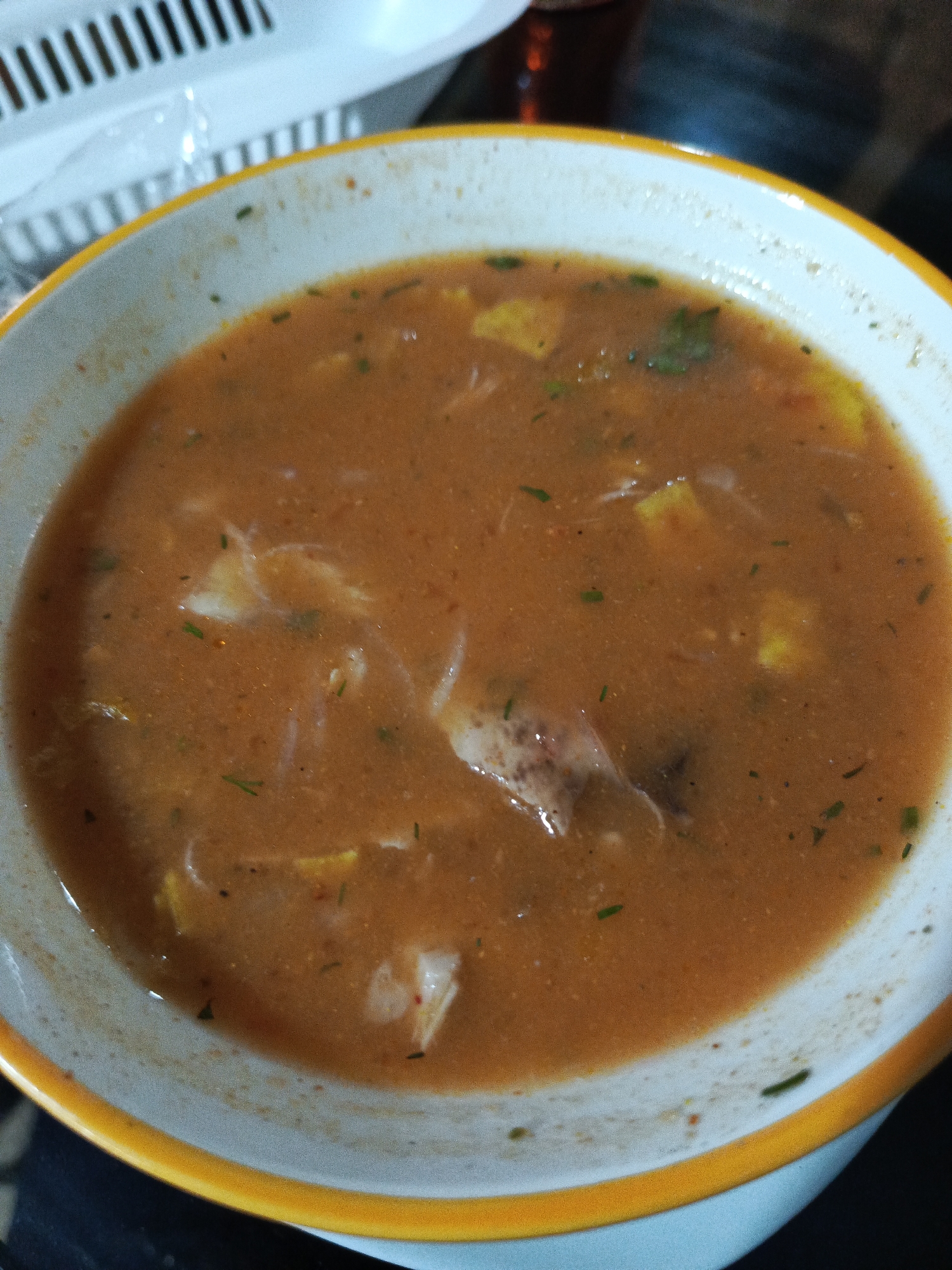 A close-up, top-down shot shows a bowl of hearty soup. The soup is a reddish-brown color with visible chunks of meat and pieces of what appears to be vegetables. Small green herbs are sprinkled throughout the broth. The bowl itself is white with a thick yellow rim. The background is blurred but suggests an indoor setting, possibly a restaurant. There is no visible indication of the specific location within Guayaquil, Ecuador.