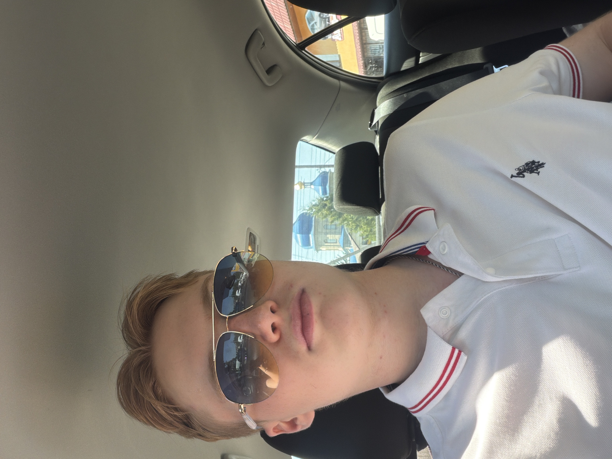 A young man with blond hair and sunglasses is sitting in the backseat of a car. He is wearing a white polo shirt with red and blue trim. The car's interior is visible, including the seatbelts and headrests. Outside the car windows, a blue-domed building, possibly a church, and other structures are visible. The scene appears to be in a city, possibly Polonne, Ukraine, given the architectural style of the building and the general appearance of the surroundings. The lighting suggests it is daytime.