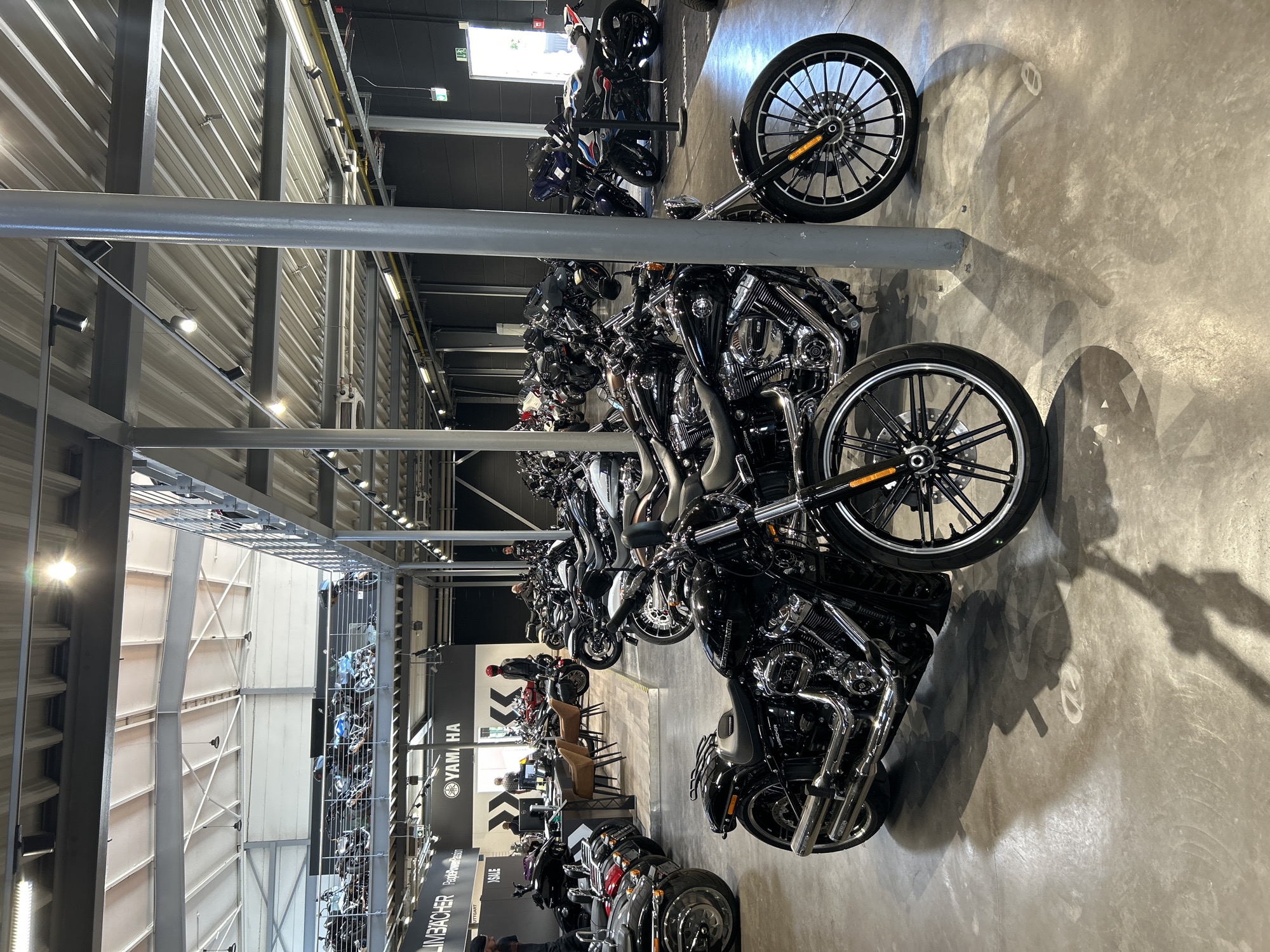 The image shows the interior of a motorcycle dealership, likely in Bernhausen, Germany. Rows of motorcycles are neatly arranged in the space, with various models and colors on display. Overhead, the ceiling structure is visible, with supporting columns and lighting fixtures. In the background, a Yamaha sign is partially visible. People are present, though not prominently featured. The floor appears to be concrete.