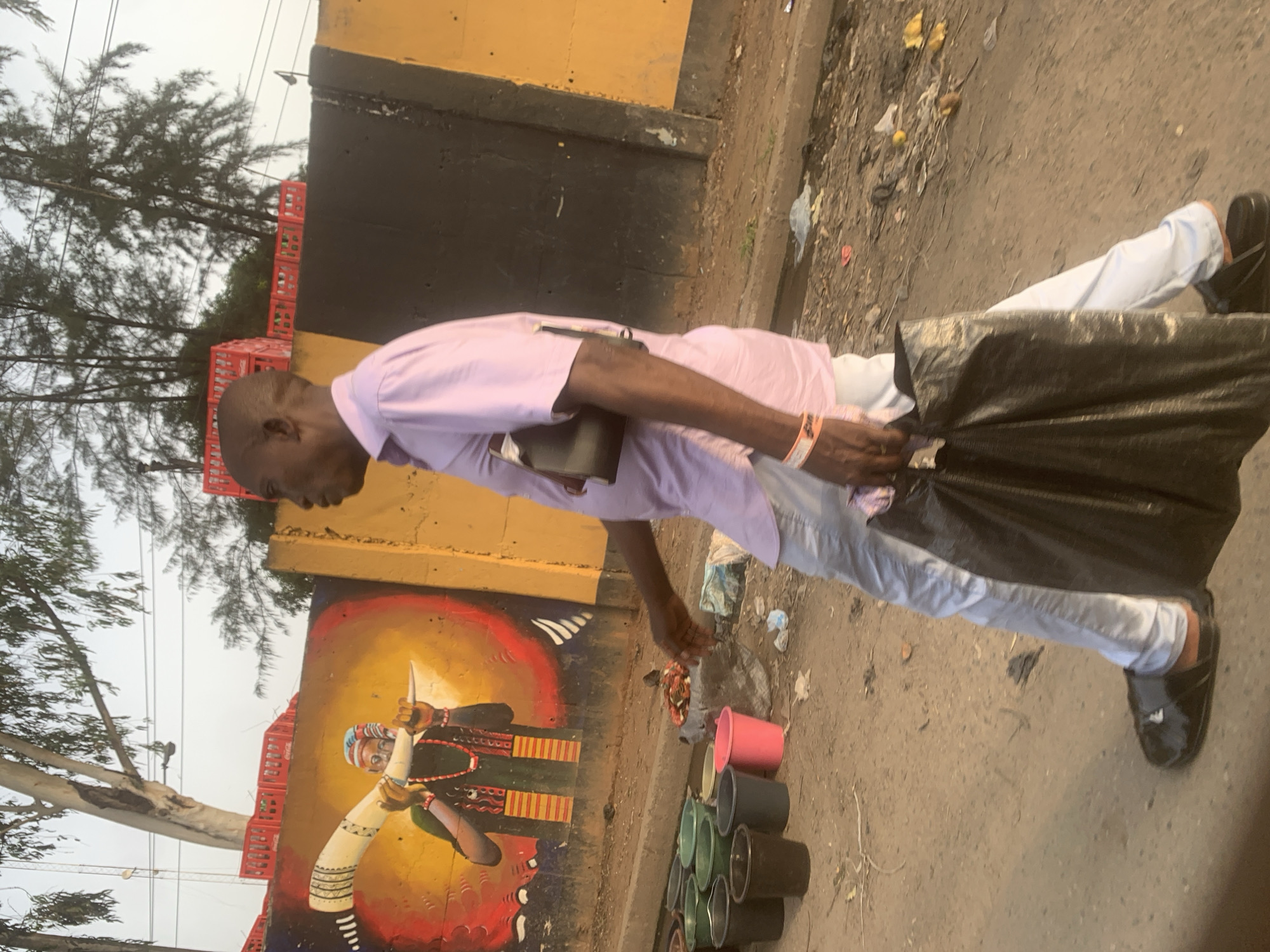 An outdoor, slightly tilted, medium shot captures a man on a dirt path next to a colorful mural and various street items. The man, seen in profile, is of average build with very short or bald hair, wearing a light purple long-sleeved shirt and light-colored trousers. He has a small black bag slung across his right shoulder and is holding a large black plastic bag with his left hand. His right hand is reaching towards a collection of small, colorful plastic buckets or pots (pink, green, silver/grey) arranged on the ground in front of him, suggesting he might be a street vendor or arranging goods.
Behind the man is a vibrant mural painted on a wall, depicting a person in traditional attire, possibly a chief or tribal figure, holding a large white horn-like object. The mural features bold red, yellow, and black colors. Above the mural and to its left, several red plastic crates, some bearing the "Coca-Cola" logo, are stacked. The ground is a mix of dirt and concrete, with some scattered debris. In the background, tall trees with sparse, needle-like foliage are visible against a hazy or overcast sky. A yellow and black striped barrier runs along the top of the wall behind the man. The scene appears to be taking place during daytime in an urban or semi-urban environment, consistent with the Fegge, Nigeria context.