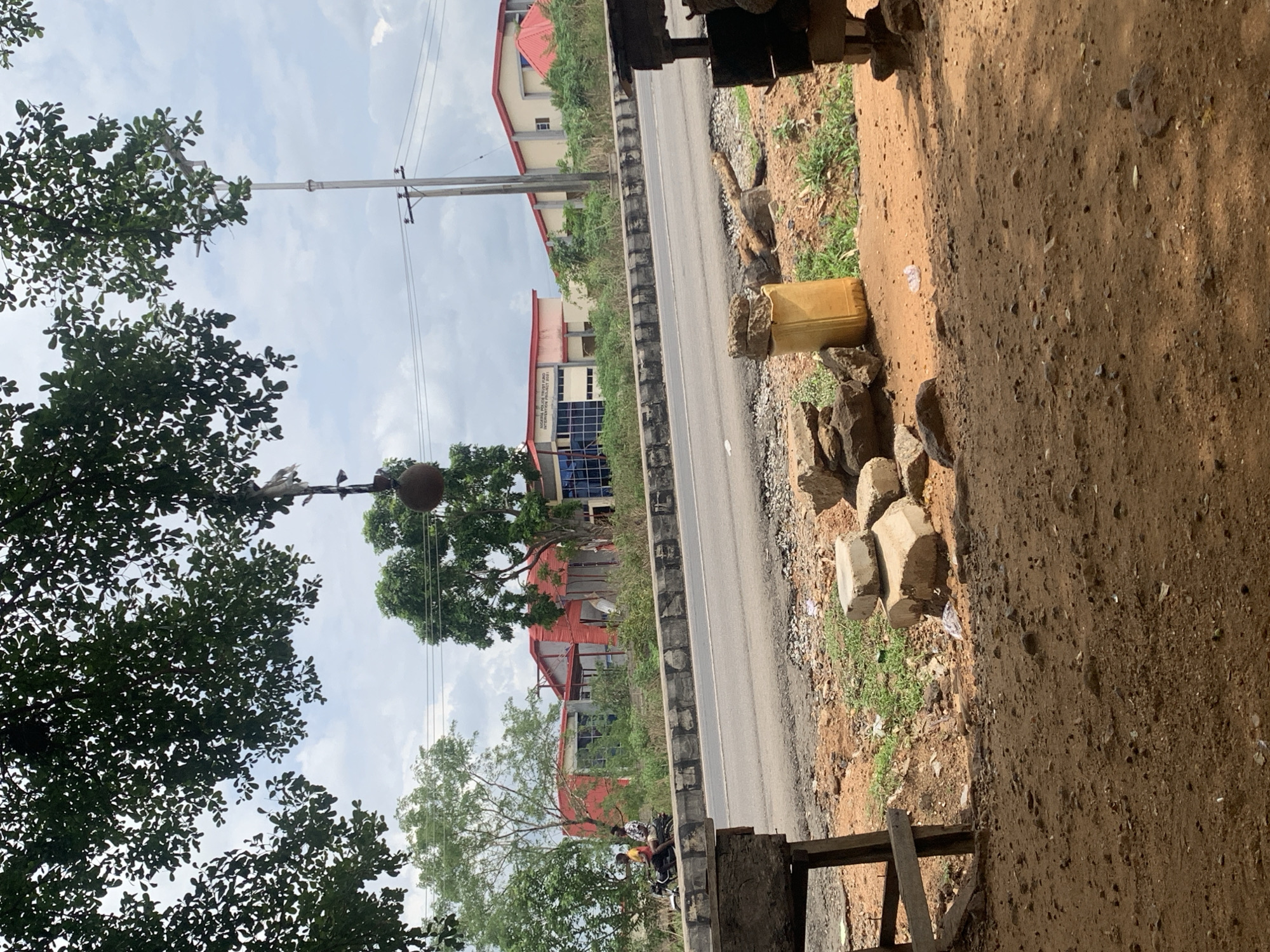 The image depicts a street in Lokoja, Nigeria. The street is lined with buildings, some with red roofs. Power lines are visible above. There are trees on both sides of the road. Two people are riding a motorcycle on the road. Construction materials such as rocks, yellow containers, and concrete blocks are near the edge of the road, on the right side of the frame. The ground is dirt.