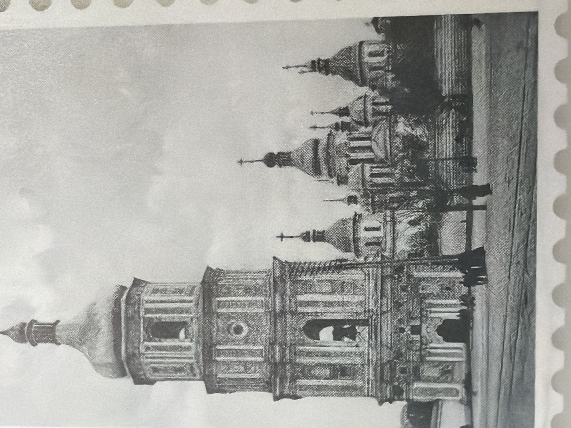 The image is a black and white photograph of the Bell Tower of St. Sophia Cathedral in Kyiv, Ukraine. The Bell Tower is a tall, multi-tiered structure with a distinctive golden dome. Adjacent to the tower, to the right, are the domes and spires of St. Sophia Cathedral itself, with multiple onion-shaped domes topped with crosses. In the foreground, there is a paved area with what appear to be tram tracks. A few silhouetted figures of people are visible near the entrance of the cathedral complex, and some bare trees are scattered in the middle ground, suggesting a cool season. The sky above is overcast. The image is framed by a perforated edge, characteristic of a postage stamp.