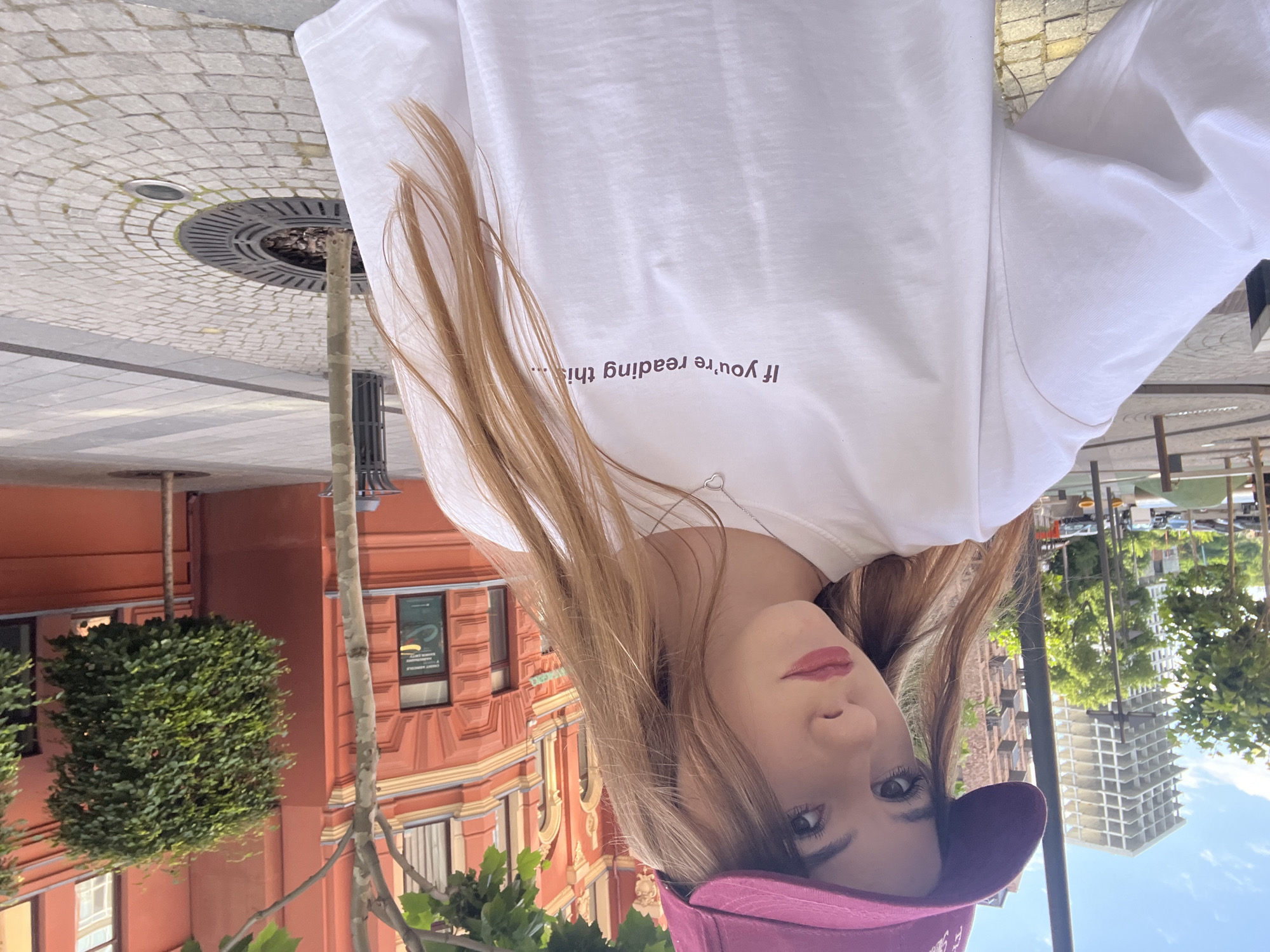 A young woman with long, light brown hair and dark eyes, wearing a maroon baseball cap and a white t-shirt with the text "If you're reading this..." printed on it, poses for a selfie outdoors in Dnipro, Ukraine. The background shows an urban environment with a modern building under construction, an orange building with arched windows, trees, and a paved walkway with cobblestone patterns. The woman has light makeup and a necklace with a heart-shaped pendant. The lighting suggests it is daytime.