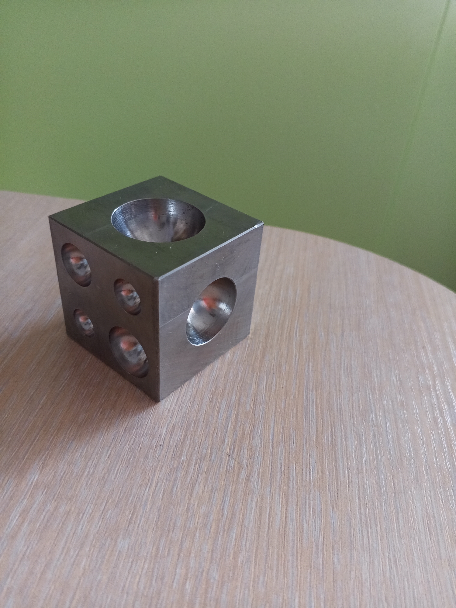 A metallic cube with several hemispherical indentations on its faces rests on a light-colored wooden surface. The cube is positioned in the foreground, slightly to the left of the center of the image. Behind the cube, a solid green wall serves as the background. The lighting suggests an indoor environment, possibly a room. The image is a still photograph with no discernible activity or interaction taking place. The exact location in Vilyuysk, Russia, cannot be determined from the visual information provided.