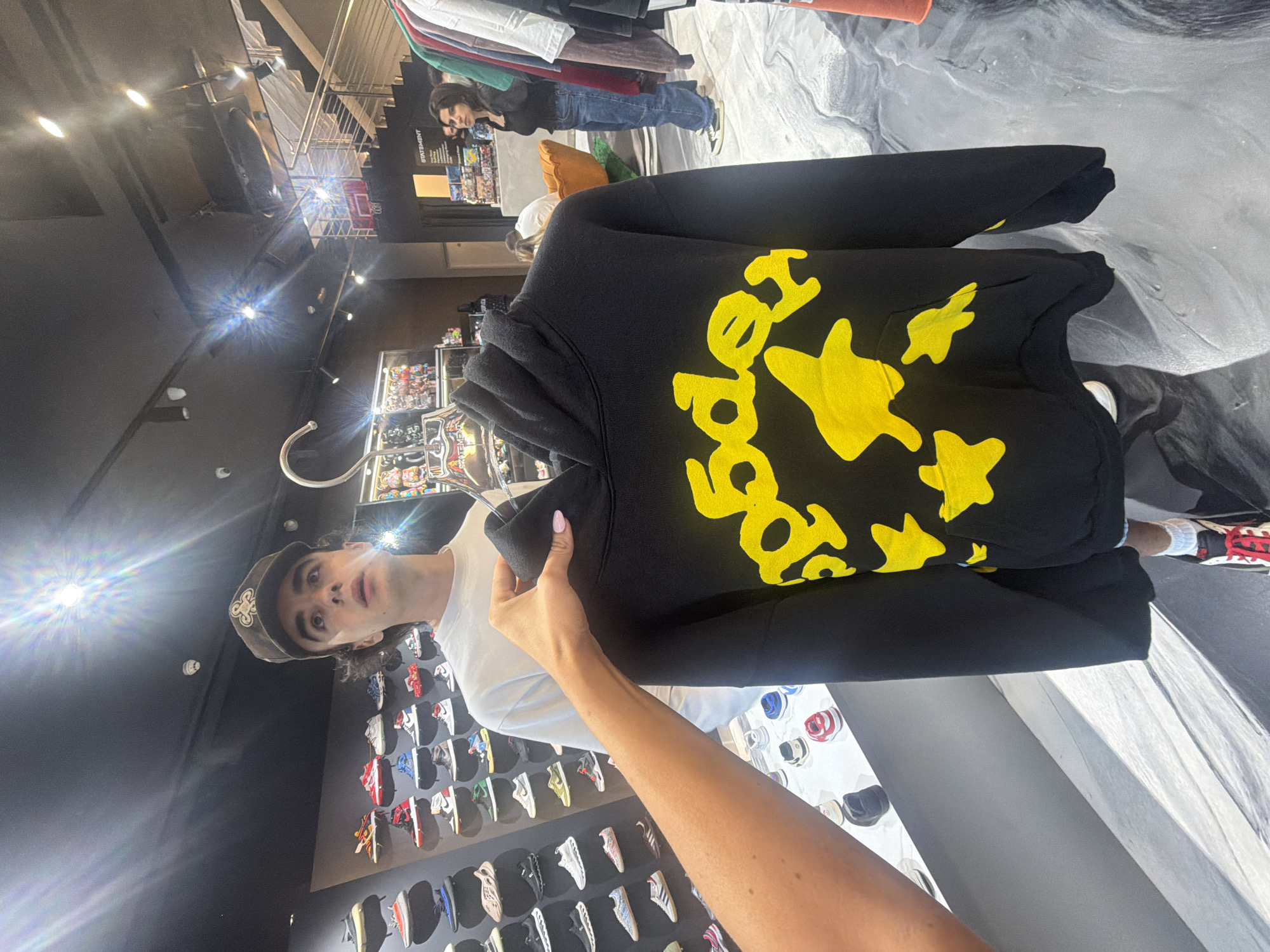 A young person wearing a white t-shirt and a brown baseball cap with a logo is standing in a sneaker store, looking upwards. A hand is holding up a black hoodie with a yellow graphic design to the camera, obscuring the upper torso of the person. The hoodie features a yellow splatter-like design and what appear to be stars. In the background, shelves are stacked with various sneakers. Further back, a young woman in blue jeans and a black top is browsing clothes on a rack, and another person is partially visible sitting on an orange chair. The store appears to be well-lit with overhead lighting and natural light filtering from an upper level. The environment suggests a retail space, likely a sneaker boutique, potentially in London, United Kingdom, given the context of such establishments and the presence of fashion-conscious individuals.