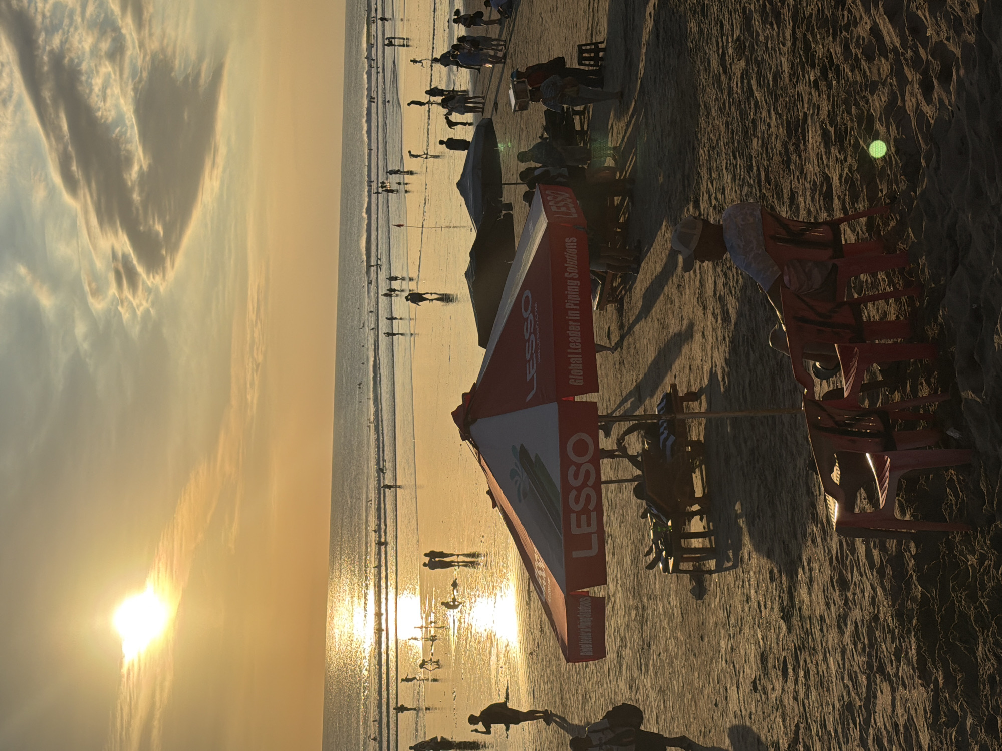 This media file captures a vibrant beach scene in Kuta, Indonesia, during sunset. The golden hues of the setting sun cast a warm glow across the sand and sea, creating a serene and picturesque atmosphere. Silhouettes of numerous people dot the shoreline, some strolling along the water's edge, while others gather in small groups, enjoying the tranquil evening.
The foreground features beach chairs and umbrellas, hinting at a recreational area. One prominent umbrella displays bold red and white branding with visible text: "LESSO Global Leader in Piping Solutions". The sandy terrain shows textures from the receding tide, with small waves gently lapping the shore.
In the distance, the vast ocean stretches towards the horizon, reflecting the sunset's brilliance. The sky is adorned with wispy clouds, also illuminated by the sun's descent. The overall mood is peaceful and relaxed, with the gentle sounds of the waves and the warm glow of the sunset contributing to the idyllic ambiance. The presence of numerous people enjoying the beach suggests a popular tourist destination, characteristic of Kuta.