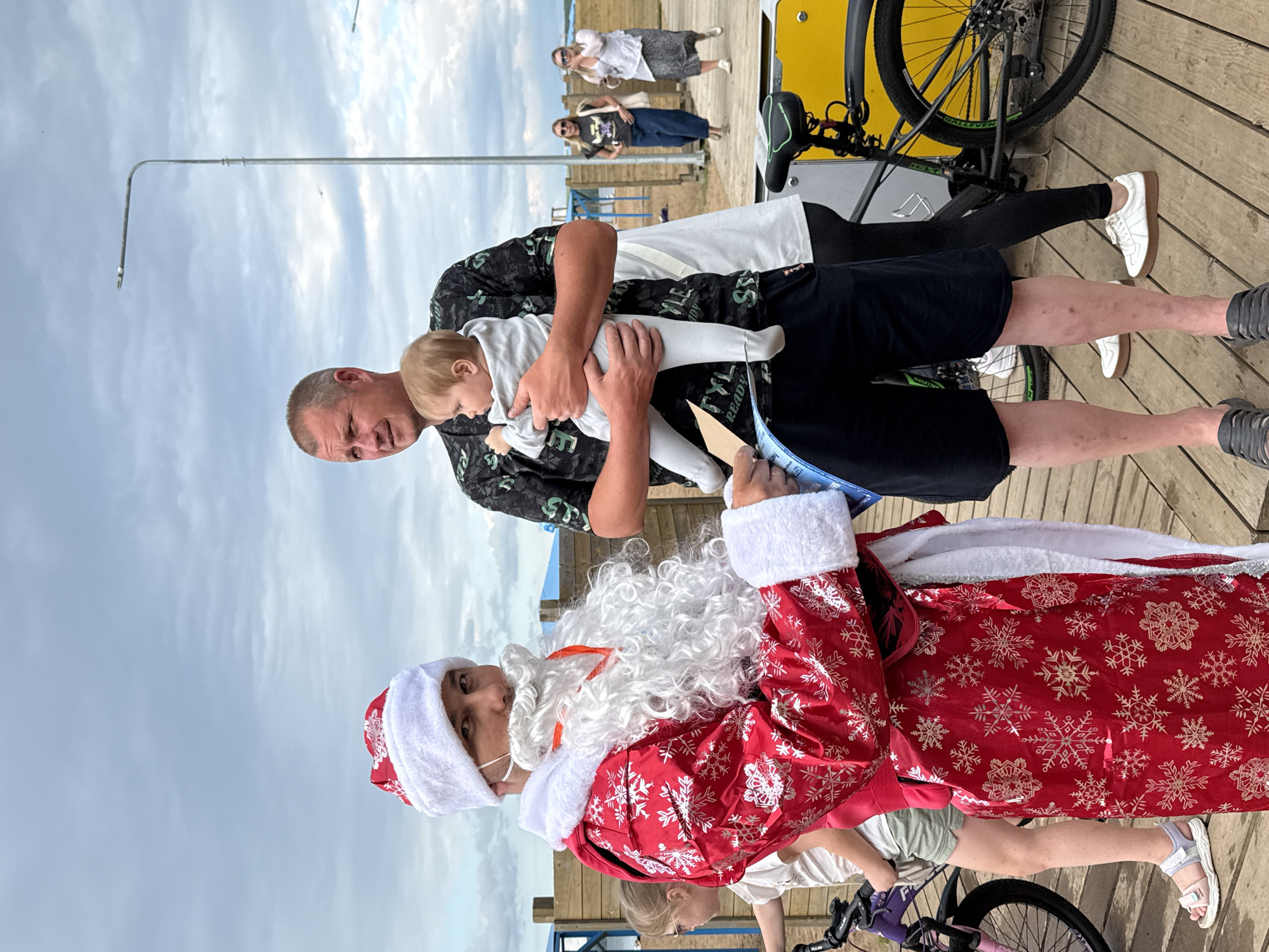 In Chaikovsky, Russia, on a wooden boardwalk near a body of water, a man dressed as Santa Claus stands facing the camera. He is wearing a red Santa suit with white trim and snowflake patterns, a white beard, and a face mask. He is holding a brochure or pamphlet. To his right, a man holds a baby dressed in a grey onesie. In the background, a woman wearing a white top and patterned skirt walks away from the camera, followed by another woman. Several bicycles are visible, including one with a yellow frame. The sky is overcast, suggesting a cloudy day.