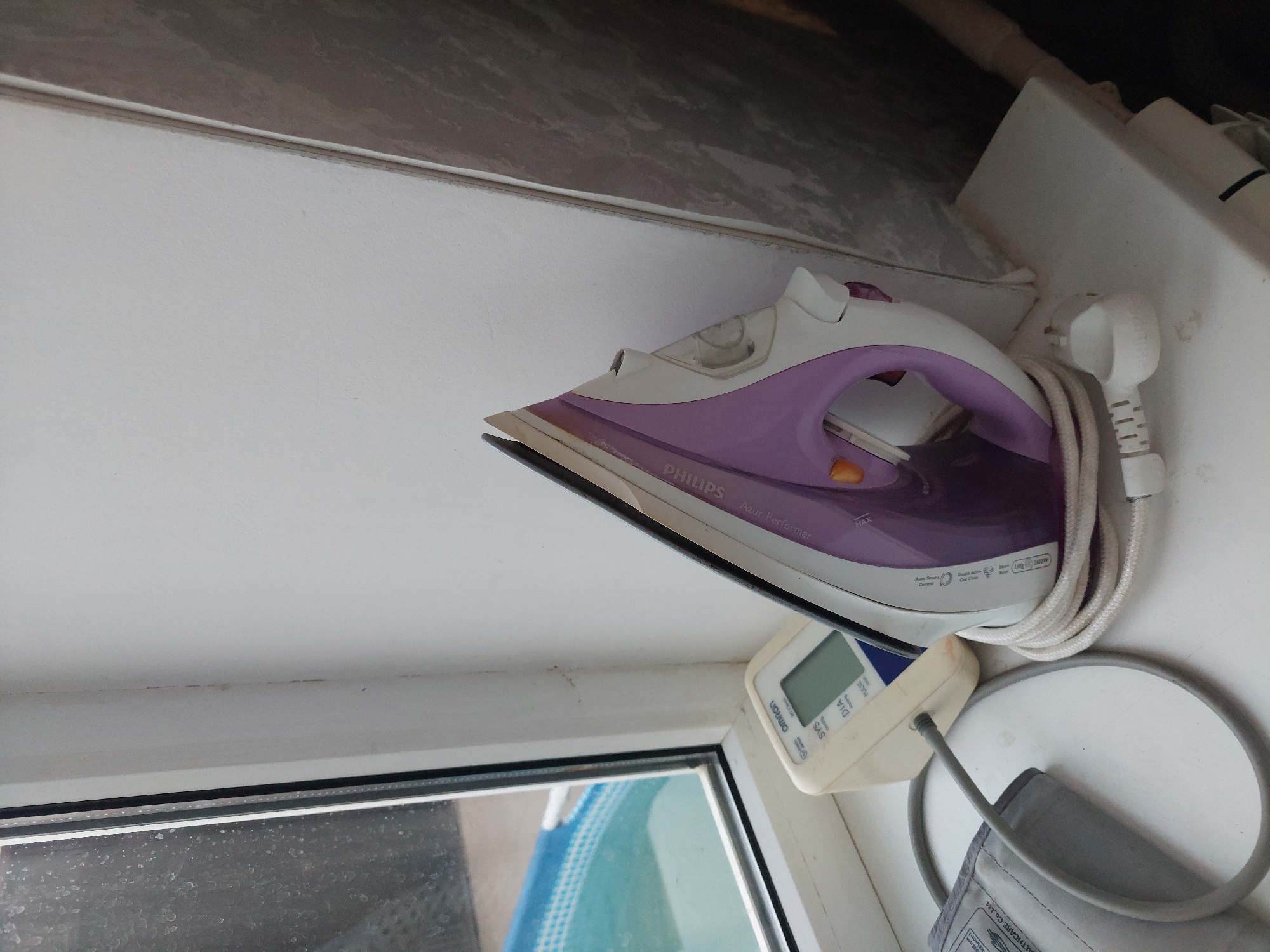 The image depicts a domestic scene on a windowsill in Makhachkala, Russia. A white and purple Philips Azur Performer steam iron rests on the windowsill, its power cord neatly wound around its base. Next to the iron is a white Omron blood pressure monitor with a digital display and a connected cuff. In the background, a window offers a view of an inflatable swimming pool with blue patterned edges. The windowsill itself is white, and the wall behind it is also white, with some minor discoloration and dust visible. The overall impression is one of everyday household items stored in a residential setting.