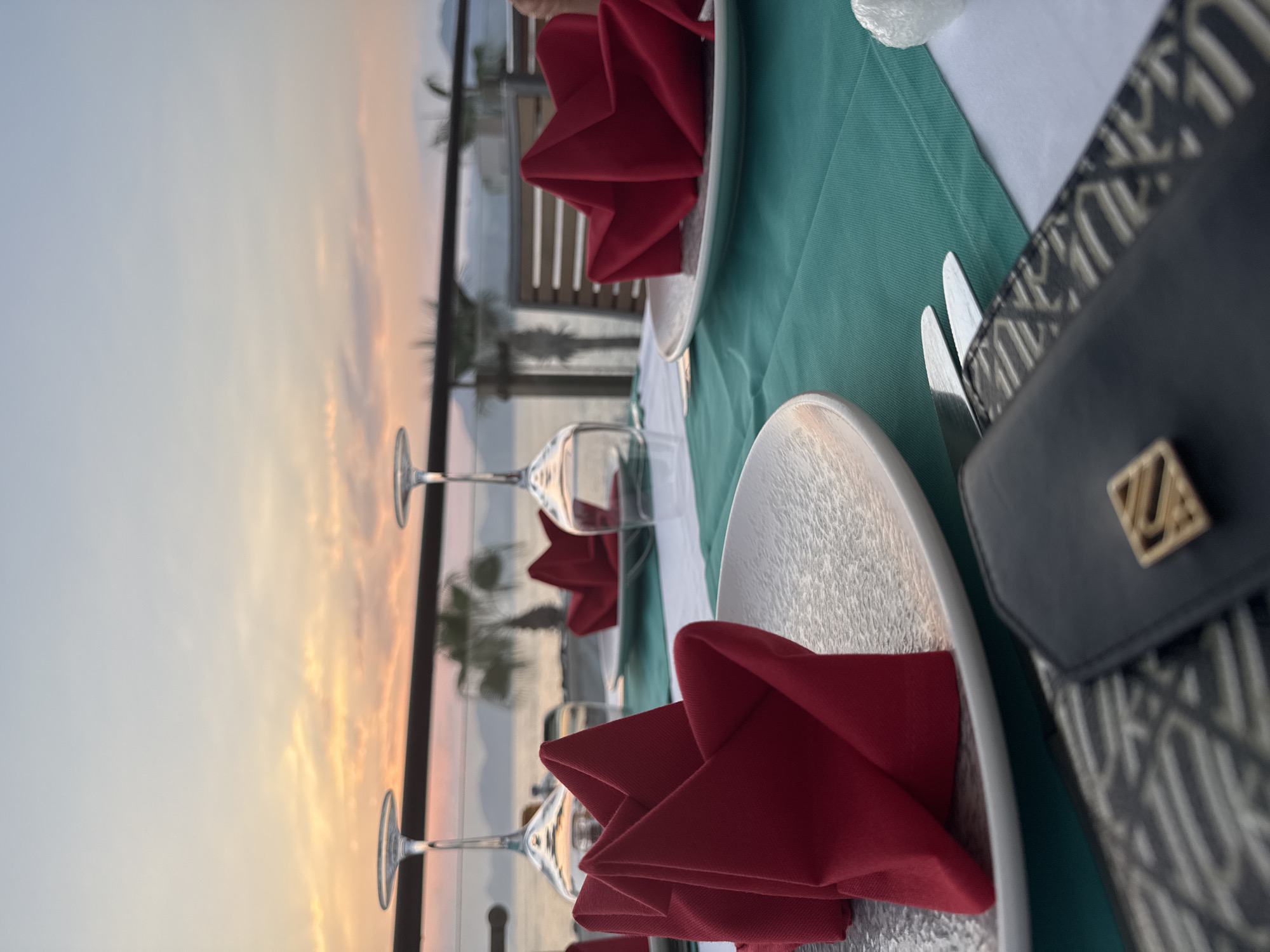 An outdoor dining table is set for a meal at sunset, with a view of the sea and mountains in Fethiye, Turkey. The table is covered with a white tablecloth and a teal runner. There are two white plates, each with a red, folded napkin. Two wine glasses are visible, one filled with liquid. In the foreground, a black clutch with a gold emblem rests on a patterned surface. Palm trees are visible in the background. The sky is filled with the warm colors of sunset, with clouds tinged with orange and pink. The sea is calm and reflects the colors of the sky. The overall atmosphere is serene and romantic.