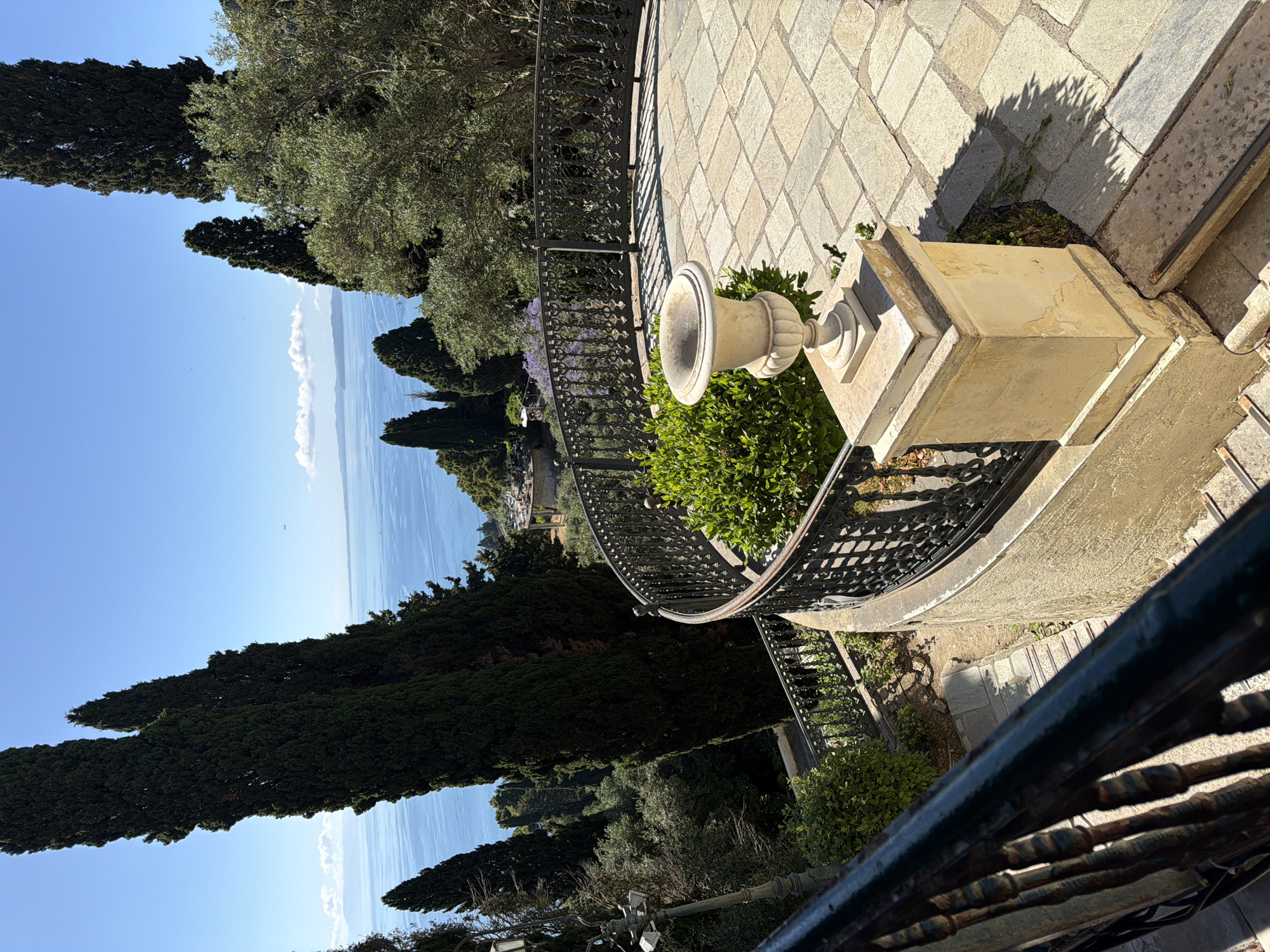 An expansive, sunlit view from a stone terrace overlooks a vast blue sea, set in Gastouri, Greece. The foreground features a light-colored stone-paved terrace with an ornate black metal railing. A prominent classical stone urn rests on a square pedestal amidst lush green bushes to the right, casting clear shadows on the paving.
Beyond the railing, the mid-ground is dominated by dense Mediterranean vegetation, including tall, slender cypress trees and olive trees with lighter green foliage. These trees frame the panoramic vista of the water.
In the background, a calm, deep blue sea stretches horizontally to meet a bright, clear blue sky. A few wispy white clouds are scattered along the distant horizon over the sea, and a faint landmass is visible in the far distance across the water. The weather is sunny and clear, suggesting daytime, likely morning or early afternoon given the angle of the shadows. No people are visible, and no activity is explicitly taking place, lending a tranquil and serene atmosphere to the scene. There is no visible text.