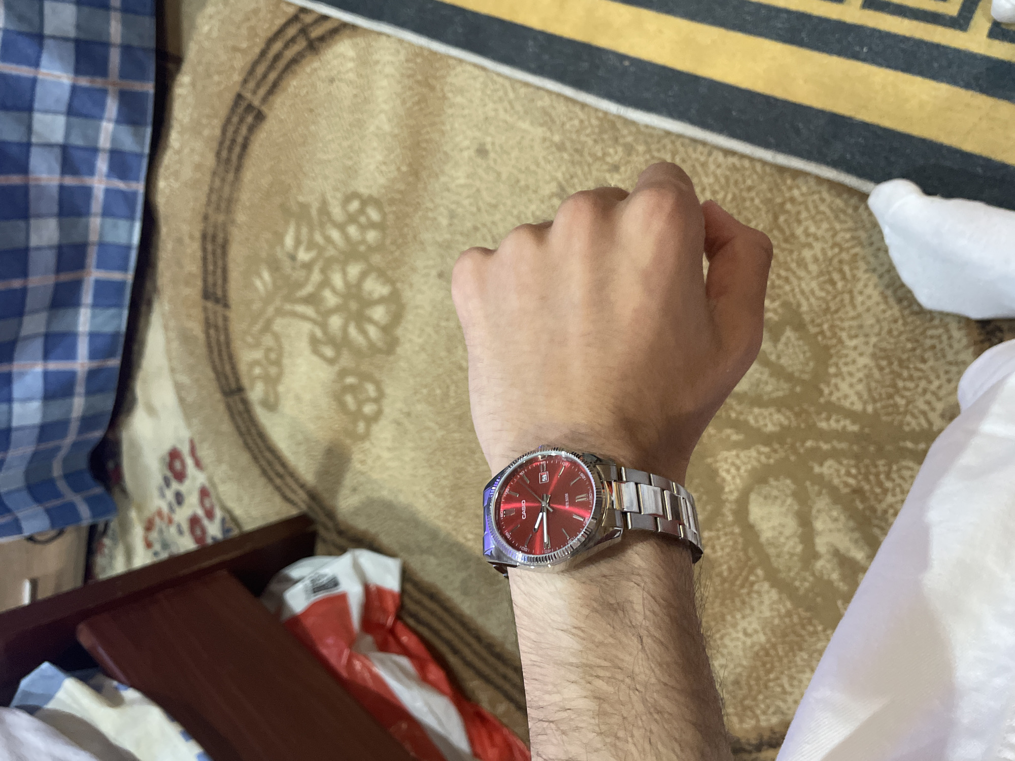 An overhead shot captures a person's left arm and hand, adorned with a silver-toned wristwatch, against a backdrop of a patterned rug. The scene is set indoors, within what appears to be a room in Ihtiman, Bulgaria.
The central subject is the wristwatch, featuring a striking red dial, silver-tone stainless steel bracelet, and a fluted bezel. The watch shows a date "31" in a window at the 3 o'clock position. The brand "CASIO" is clearly visible on the watch face, along with the text "WATER RESIST". The person's hand is slightly clenched.
Surrounding the arm are various elements that form the indoor setting. A patterned rug, primarily in shades of beige and brown with decorative motifs, covers the floor. Parts of other objects are visible: a blue and white checkered fabric (possibly a shirt or blanket) is to the left, and a white garment, perhaps a traditional thobe or a casual piece of clothing, is to the right. In the lower left quadrant, a dark wooden edge, possibly part of furniture like a bed frame or a drawer, is seen, beneath which lies a crumpled red and white object, resembling a plastic bag.
The lighting suggests an indoor environment, but the specific time of day or external weather conditions cannot be determined. No particular activity is shown beyond wearing the watch; the scene appears to be a casual, static moment.