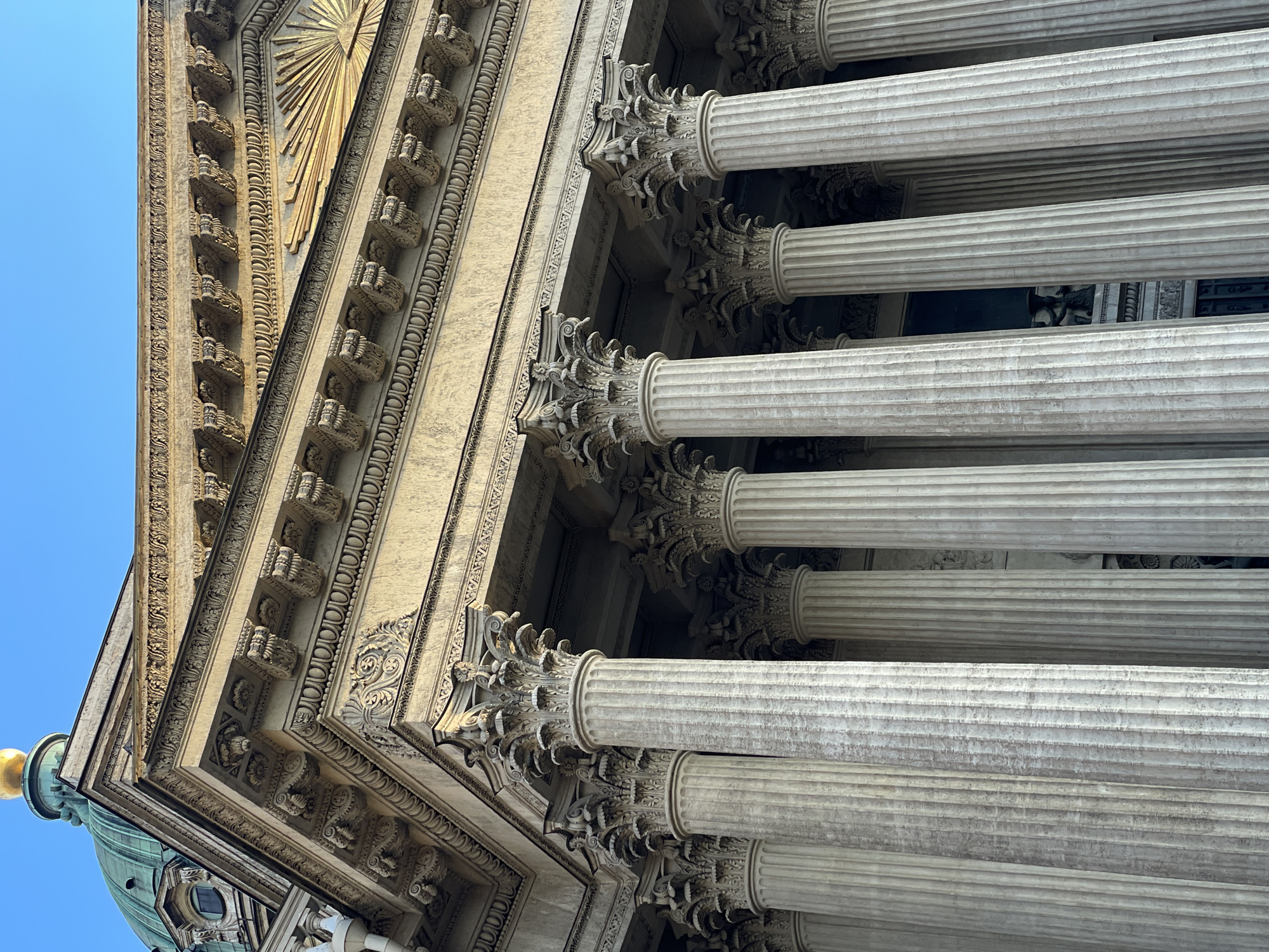 An upward-angled view captures the imposing neoclassical architecture of a grand building in Saint Petersburg, Russia. The perspective emphasizes the towering height and intricate details of the facade.
Several massive, light-colored fluted columns with highly ornate Corinthian capitals dominate the right side of the frame. These capitals are intricately carved with acanthus leaves and other decorative motifs. The columns extend vertically, appearing to support a heavy entablature and cornice, which is also richly adorned with repetitive patterns and sculpted elements.
The main facade of the building, visible on the left and center, features detailed cornices and friezes with continuous decorative moldings. A prominent golden, sunburst-like emblem with radiating lines is positioned on the upper section of the facade, shining brightly. A partial view of a green-patinated dome, topped with a golden finial or cupola, is visible at the lower-left corner of the image, hinting at the larger structure of the building, possibly a cathedral or a significant public edifice.
The scene is set against a clear, vibrant blue sky, indicating bright daylight conditions. The strong sunlight highlights the architectural details and creates distinct shadows, adding depth and texture to the stone work. There are no people visible, and no specific activity is taking place; the image focuses solely on the grandeur and craftsmanship of the architecture. No visible text is present in the image.