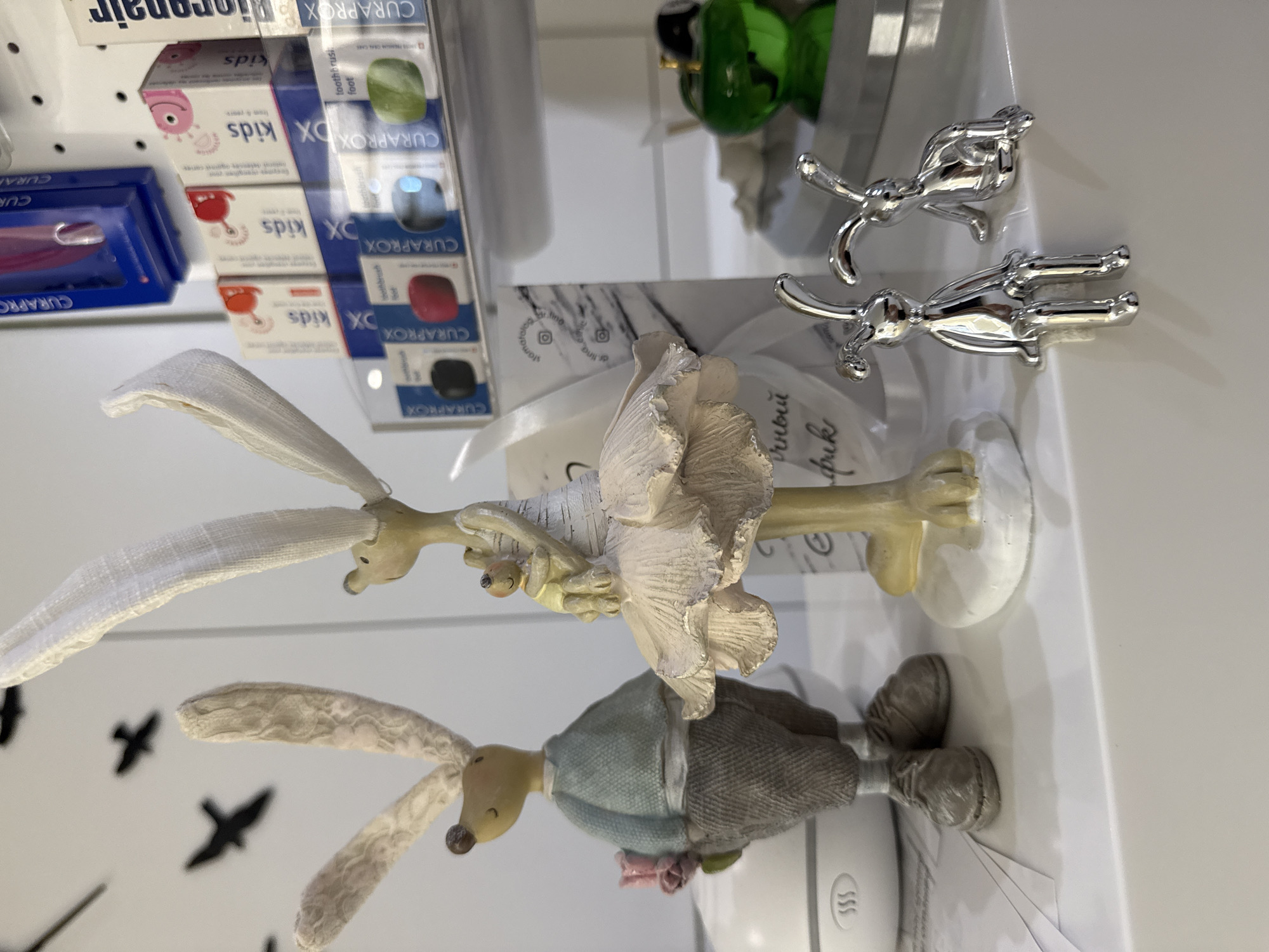 The image displays a collection of decorative items on a white shelf. In the foreground, two silver metallic rabbit figurines are positioned side-by-side, with one sitting upright and the other in a crouched position. Behind them, a larger figurine of a rabbit dressed in a light blue shirt and grey trousers, holding a pink flower, stands to the left. To the right of this figure is another rabbit figurine, appearing to be a ballerina, adorned in a white tutu and holding a smaller bunny. In the background, shelves are filled with various Curaprox dental products, including toothbrushes and toothpaste, indicated by their distinctive packaging. The overall setting appears to be a retail or display environment, potentially a pharmacy or a specialized store. There are no visible signs indicating a specific location within Мирновское сельское поселение, Ukraine.