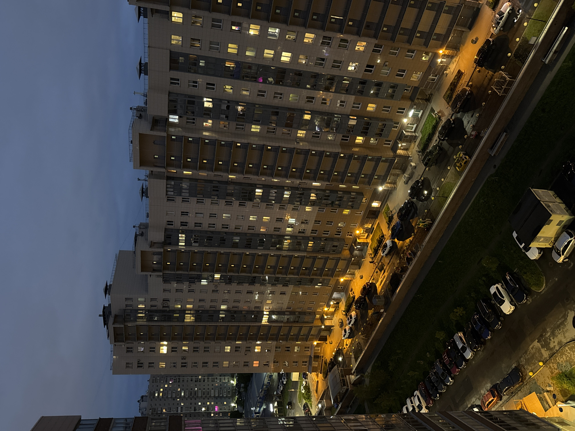 The image presents an elevated view of a densely populated urban environment, likely taken at twilight or dusk. Predominantly, the scene is occupied by a cluster of high-rise apartment buildings with numerous illuminated windows, suggesting residential occupancy. These buildings are organized in a block-like structure, with visible balconies and rooftop architectural details. The street-level infrastructure includes a road with parked cars and streetlights, as well as patches of vegetation or green spaces. The lighting from the streetlights and the building windows casts a warm, artificial glow that contrasts with the dark and fading sky above. The presence of parked vehicles and illuminated windows suggests daily activity is still ongoing. Based on architectural style and signage elements, the location is tentatively identified as being in an urban area of Russia.
