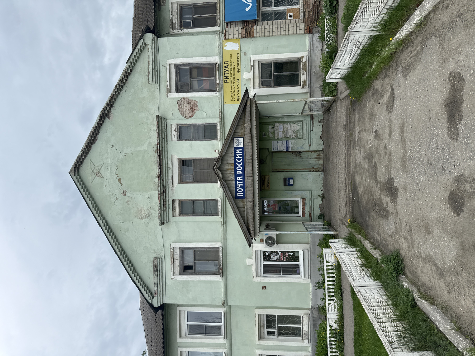 The image depicts a two-story building in Ленино, Russia. The building is painted light green and appears to be in disrepair. Above the entrance is a sign that reads "ПОЧТА РОССИИ" (Post of Russia). To the left of the entrance, a sign advertises "РИТУАЛ" (Ritual), a company providing funeral services. A sidewalk runs along the right side of the building, bordered by a low white fence. The sky is overcast.