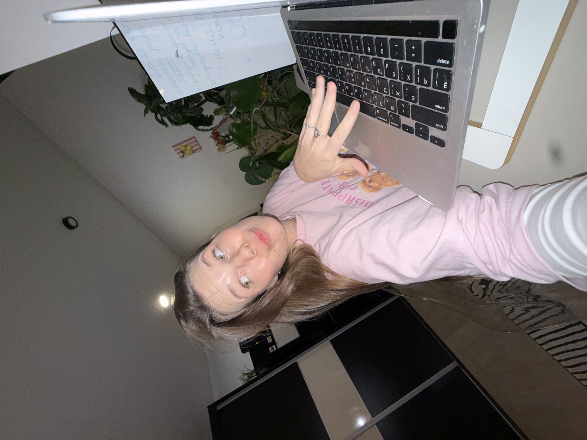 A selfie-style photo shows a person with light skin, light brown hair, and wearing a pink shirt. They are positioned in front of an open laptop, with the keyboard visible and their hand partially obscuring it. A whiteboard with handwritten notes is placed to the left of the laptop. In the background, there's a plant, a small decorative item on the wall, and a dark-colored cabinet. The image appears to be taken indoors.