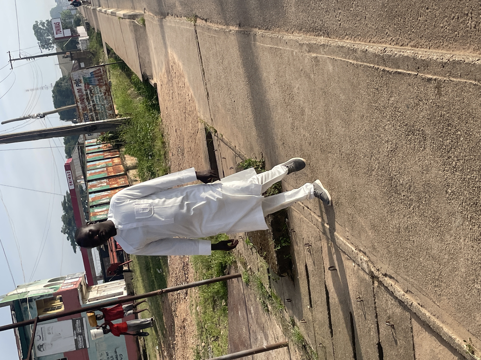 A man wearing a white, long-sleeved traditional outfit and white pants, walks towards the right of the frame on a concrete sidewalk. He has dark skin and is looking at the camera. He is wearing grey sneakers. Behind him, on the left side of the image, a building with red and green trim is visible. Two other people are in the background, one carrying a yellow bucket on their head and the other wearing a red shirt and grey pants. A large poster is attached to a pole in front of the building. Further in the background, there are rusty metal structures, some signage, and utility poles. The scene appears to be outdoors, on a street in Jos, Nigeria.