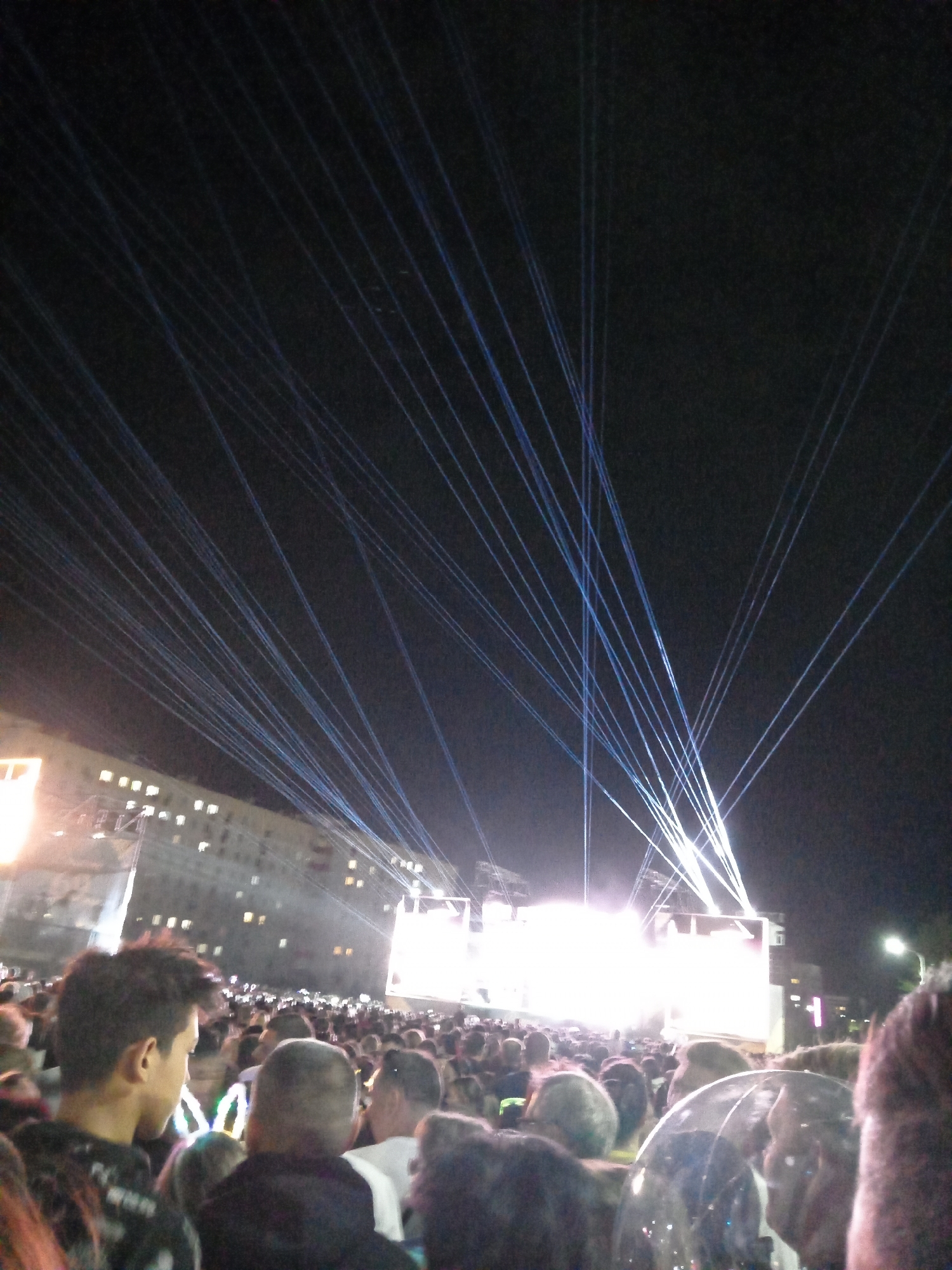 The image depicts an outdoor nighttime concert event, likely in a public space. A large crowd of people is visible, mostly with their backs to the camera, facing a brightly lit stage or platform. The stage is obscured by intense light, but appears to be a substantial structure, possibly with large screens or lighting rigs. Above the stage and crowd, numerous thin blue and white light beams, resembling laser lights, are projected upwards into the dark sky, creating a dramatic visual effect. In the background, a multi-story building with illuminated windows can be seen, suggesting an urban setting. The overall atmosphere suggests a lively event with a focus on visual spectacle. The city of Rudny, Kazakhstan, is mentioned as the location, implying this event is taking place there.