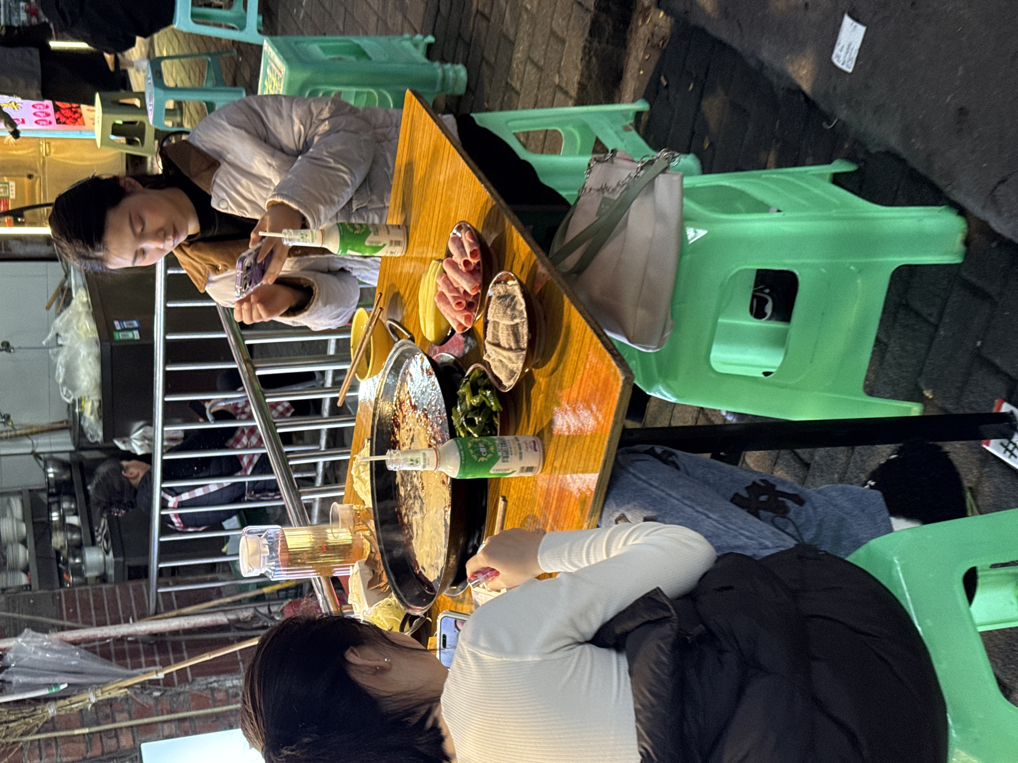 Two women are eating at an outdoor restaurant in Yuzhong District, China. One woman, wearing a light-colored jacket, sits at a table and looks at her phone. The other woman, wearing a white shirt and black jacket, is also looking at her phone. On the table are plates of food, including meat and vegetables, and bottles of sauce. Green plastic stools surround the table. The scene is lit by artificial lights. Pedestrians and street vendors are visible in the background.