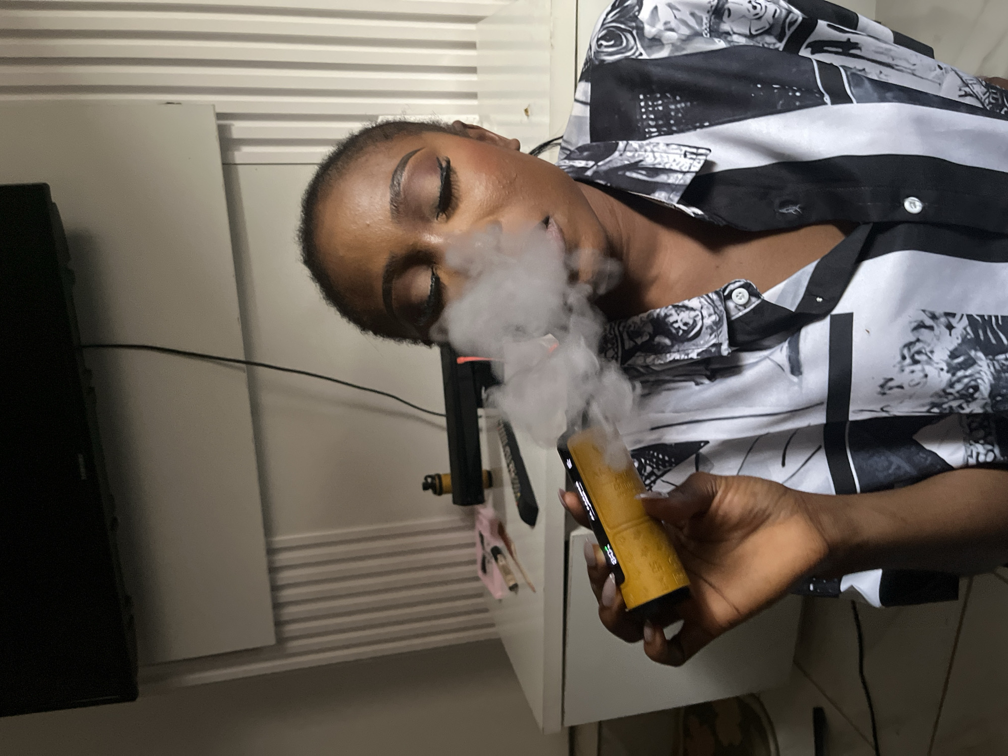 A close-up shot shows a young Black woman with her eyes closed, exhaling a cloud of smoke from a yellow vape pen. She is wearing a black and white patterned button-down shirt. The background appears to be an indoor domestic setting, with a white dresser or cabinet visible. To the left, a portion of a television screen is visible. The woman's makeup is applied, with foundation, eyeshadow, and mascara. Her fingernails are manicured. The scene is lit by artificial lighting, casting shadows. The context of the image does not provide any specific information about the location within Abuja, Nigeria.