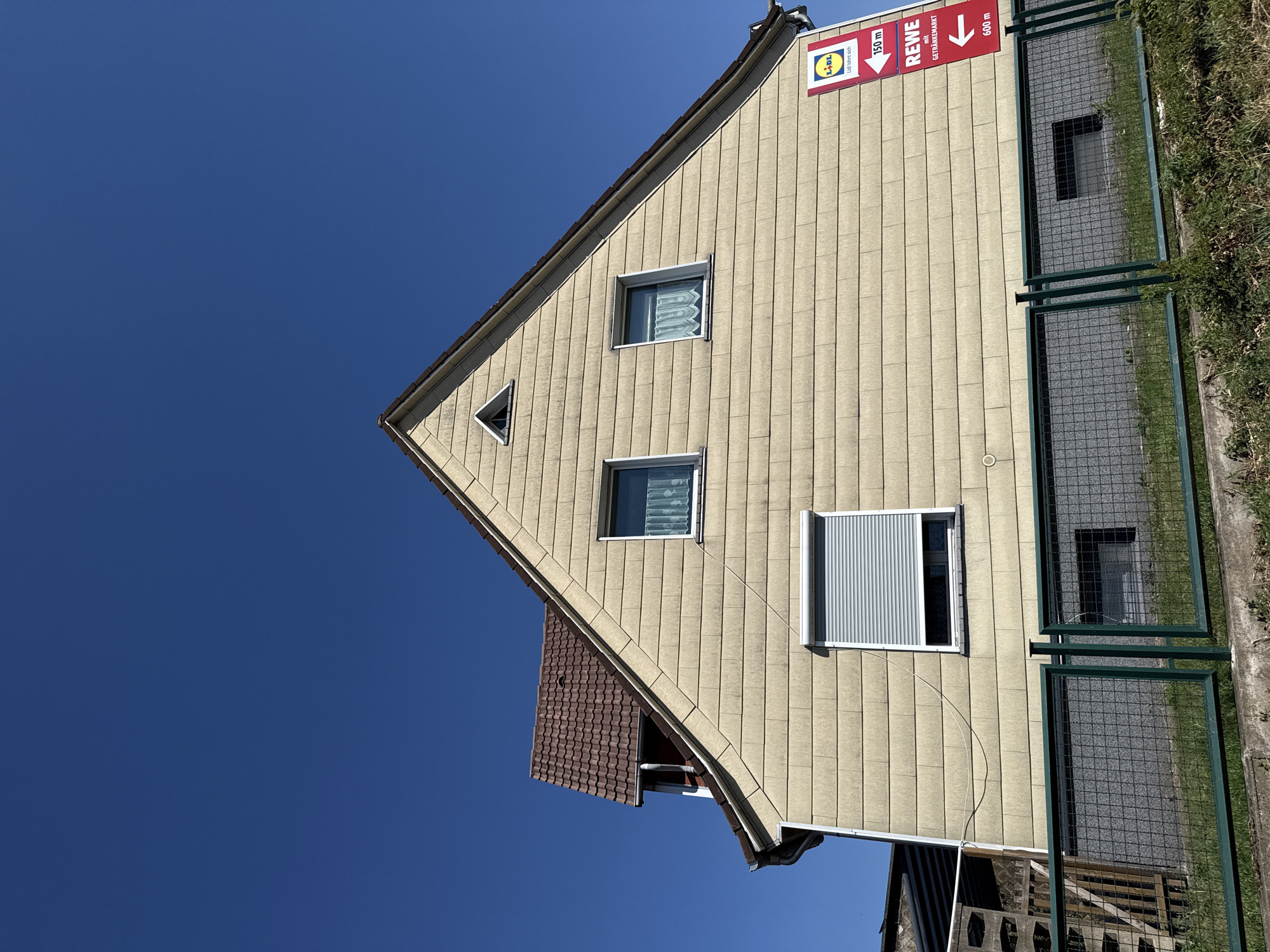 This image captures a side view of a light yellow, multi-story house against a clear blue sky. The house has a steeply pitched, brown tiled roof and several windows. One window on the upper level has a white, lace-like curtain. Another window on the lower level is covered by a grey roller shutter. A triangular window is present at the very peak of the gable.
Attached to the side of the house is a red sign with white text and logos. The top portion of the sign features the yellow and red Lidl logo with the slogan "Lidl lohnt sich". Below that, it indicates "150 m" with a white arrow pointing to the left. Further down, there is a red sign with white text that reads "REWE mit GETRÄNKEMARKT" and an arrow pointing to the left, indicating a distance of "600 m".
In the foreground, a green metal fence with wire mesh runs horizontally across the image. Behind the fence, there is a concrete foundation and a small patch of grass and green foliage. A glimpse of a darker, textured wall is visible behind the fence on the lower level of the house, with a small, dark rectangular window or vent.
The scene is set outdoors on a sunny day, indicated by the bright blue sky and lack of shadows from overhead elements. The image appears to be taken from a low angle, looking up at the house. While the specific location within Herzberg, Germany, is not explicitly stated, the presence of German store names (Lidl and Rewe) suggests it is indeed in Germany. There are no people visible in the image.