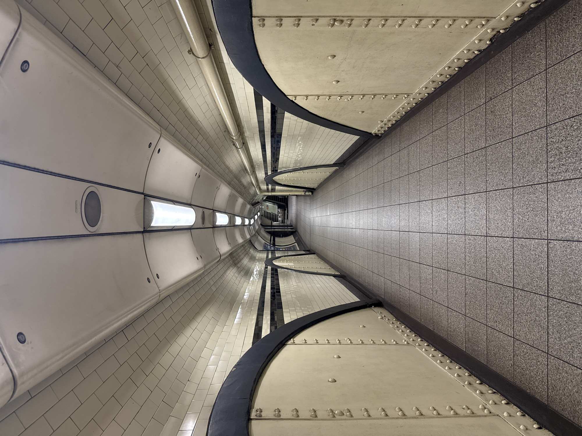 This image captures a long, curving pedestrian tunnel or walkway within an underground station, characteristic of the London Underground in the United Kingdom. The perspective is looking down the length of the passage, creating a strong sense of depth and convergence.
On the left side, the tunnel wall features curved, cream-colored panels interspersed with smaller, rectangular white tiles. These panels have circular recessed elements, possibly lights or vents, and are secured with visible fasteners. A pipe runs horizontally along the upper section of this wall. The floor on this side continues the cream and tiled aesthetic, with distinct sections of riveted panels and bold, curved black lines creating a decorative or structural border.
The right side of the passage presents a stark contrast with a flat, vertical wall covered in larger, square grey speckled tiles, suggesting a more modern or utilitarian construction. The floor on this side is also composed of these grey tiles, extending from the wall towards the center of the walkway.
The ceiling above the left side is a continuation of the tiled, curved structure, while the right side of the ceiling appears flat and cream-colored, also with visible rivets along its lower edge where it meets the grey tiled wall. The lighting is even and artificial, illuminating the passage without harsh shadows, and faint lights are visible further down the tunnel, hinting at its continuation or an exit. No people are present, lending a quiet, almost architectural study feel to the scene. There is no visible text.