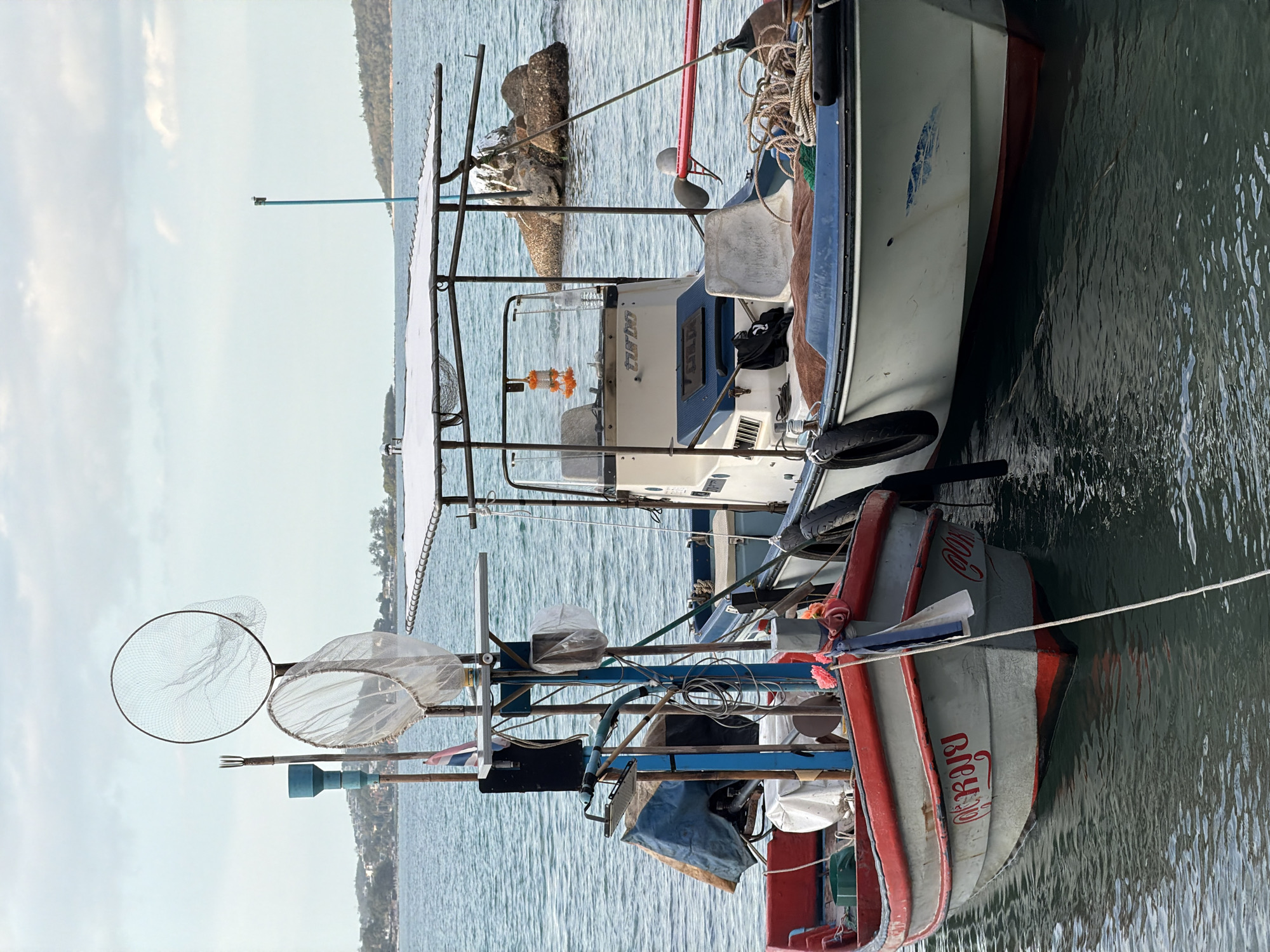 The image captures a scene of two fishing boats docked in the clear blue waters of Ko Samui District, Thailand. The foreground is dominated by the boats themselves, with the closer one, a traditional Thai fishing vessel, painted in grey with a distinctive red trim along its gunwales. Red lettering is visible on its hull. The boat behind it is a larger, white fiberglass motorboat with a canopy. Both boats are equipped with fishing gear, including nets and ropes. The surrounding environment is tranquil, with a calm sea under a partly cloudy sky. In the background, the coastline of Ko Samui is visible, characterized by lush green hills and scattered buildings. The overall impression is one of peaceful activity in a picturesque coastal setting.