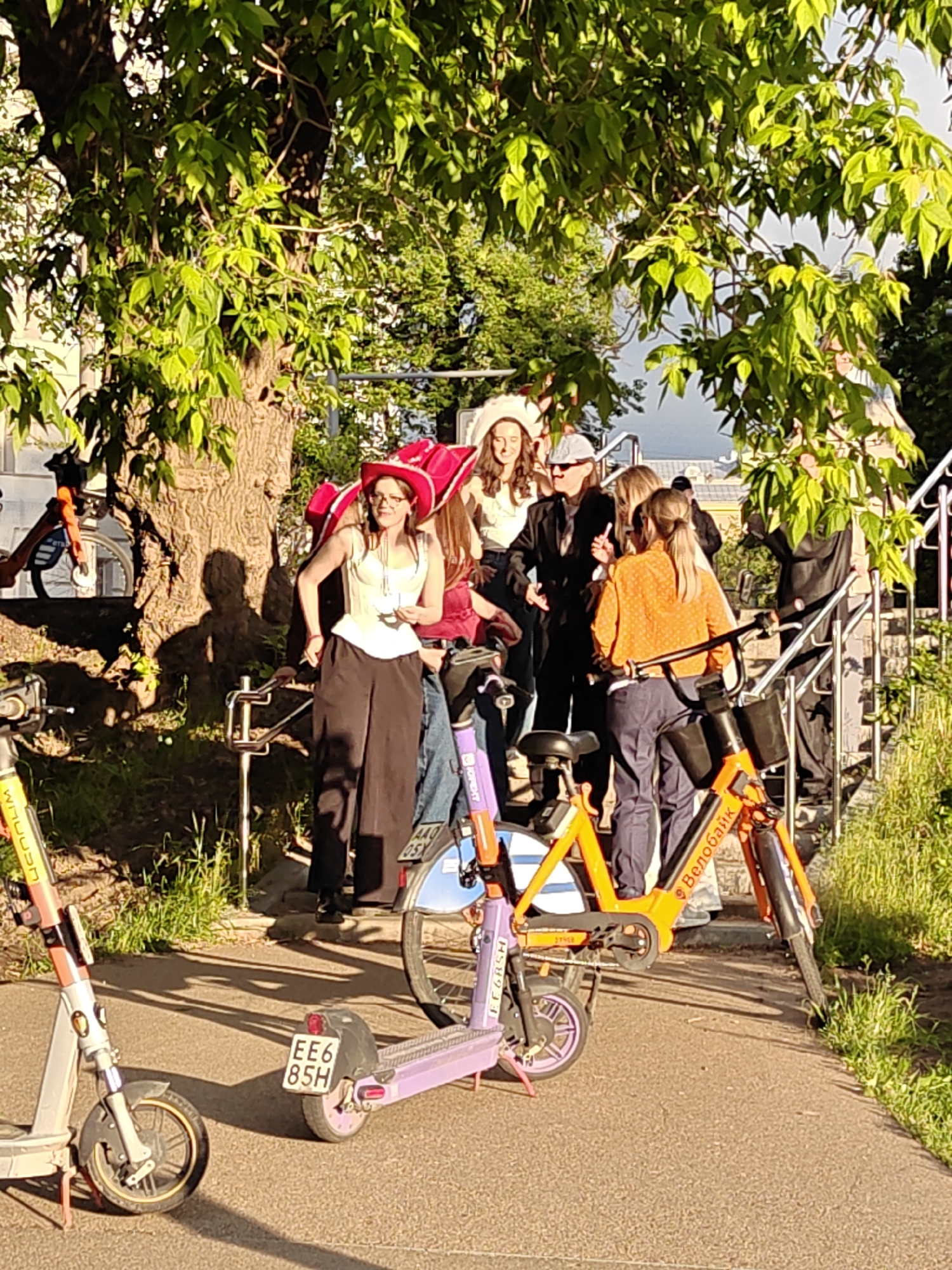 A group of people are standing on a stairway and sidewalk, accompanied by several shared electric scooters and bicycles. One scooter has a license plate that reads EE6 85H. The bikes are marked with "Велобайк," indicating the location is likely Moscow, Russia, as "Велобайк" is a popular bike-sharing service there. The people are a diverse mix of ages, genders, and attire, some wearing formal clothing and large hats. They appear to be engaged in conversation or taking a break. The scene is outdoors, with a tree providing shade and steps leading upwards in the background.