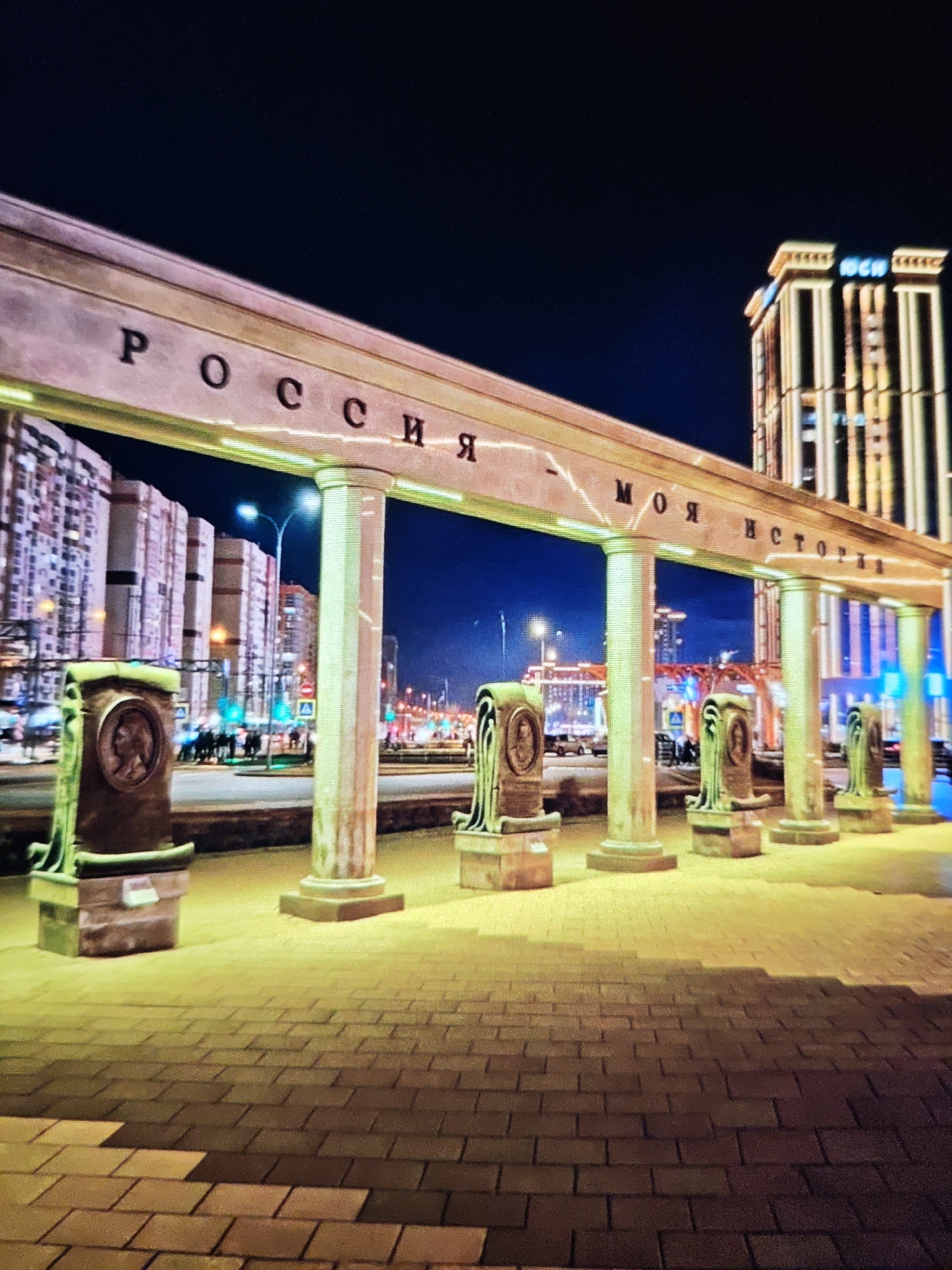 The image depicts an outdoor nighttime scene with an illuminated archway supported by columns. The inscription on the archway reads "РОССИЯ - МОЯ ИСТОРИЯ" (Russia - My History) in Russian. In front of the columns, there are several pedestals, each with a circular relief portrait. The foreground is a paved walkway with a distinct pattern of brickwork. In the background, there are multistory buildings, streetlights, traffic signals, and vehicles, suggesting an urban environment. A tall, modern skyscraper with illuminated vertical stripes and a sign reading "НСН" (likely an abbreviation for a company or organization) stands out in the background. The lighting is artificial and dramatic, casting a yellowish glow on the walkway and archway, with the sky appearing dark blue.
This particular monument is part of the "Russia - My History" museum complex. The specific location shown in the image is in Stavropol, Russia. The monument features classical architectural elements with modern lighting. The relief portraits on the pedestals are likely depictions of significant figures in Russian history. The overall scene conveys a sense of historical significance and civic pride, presented in a contemporary urban setting at night.