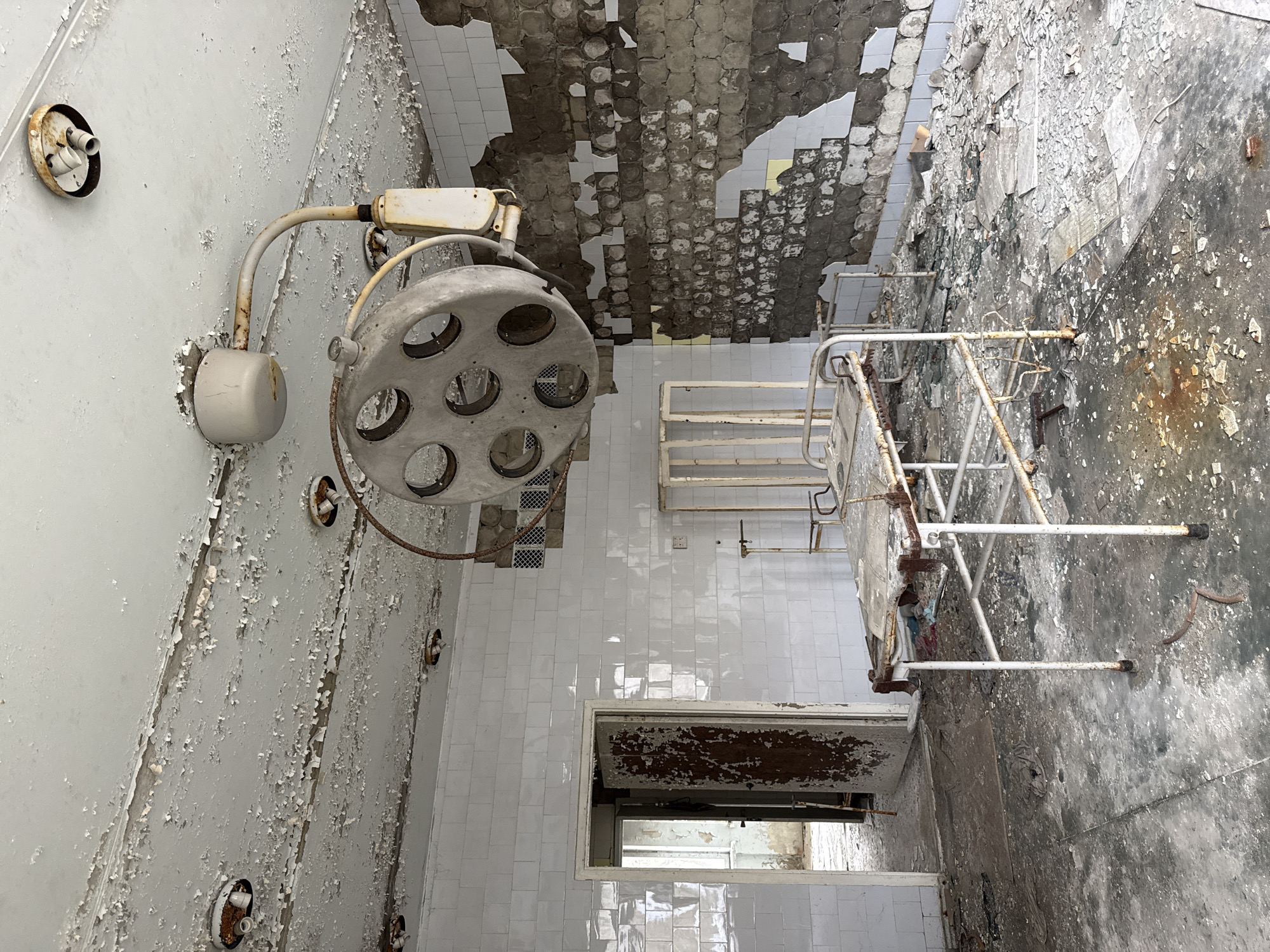 The image depicts an abandoned hospital room in Prypiat, Ukraine. Paint is peeling from the walls, and the ceiling has collapsed in sections, exposing tiles. A surgical lamp hangs in the corner, with its metal components showing rust and wear. A hospital bed frame stands in the center of the room. The floor is covered in debris, with a mix of intact tiles and areas where the surface has deteriorated. A doorway is visible in the background. The overall state of the room indicates neglect and decay.