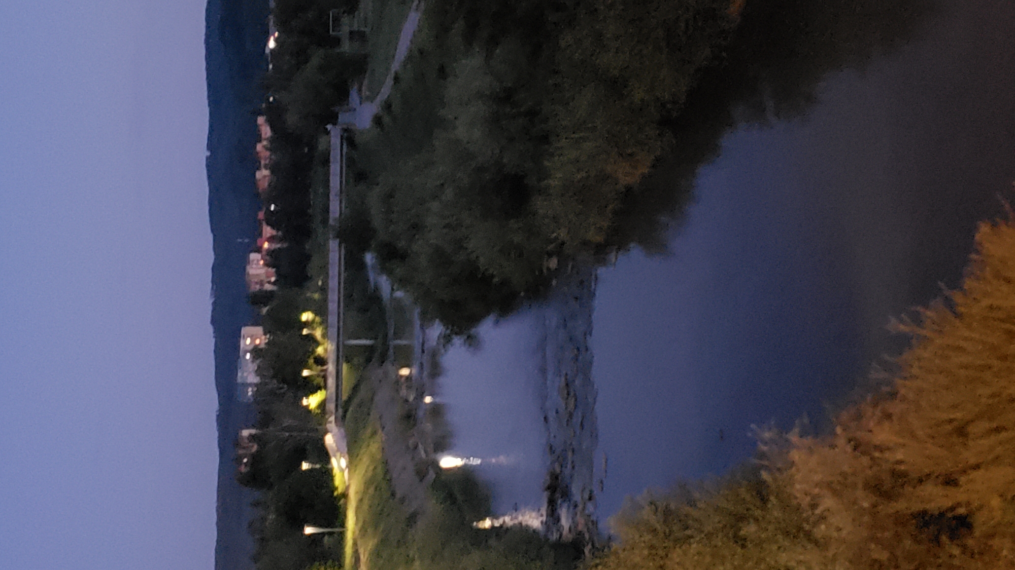 The image depicts a twilight scene in Prešov, Slovakia, likely near the Sekčov district, characterized by a calm river flowing through a park-like setting. The sky is a deep dusky blue, transitioning into darkness. On the left bank of the river, a well-lit pathway with modern lampposts is visible, suggesting a recreational area. Further back, a series of apartment buildings and houses are illuminated, casting warm light onto the twilight landscape. The river itself is dark and reflects the ambient light and distant lights from the city. Dense green foliage lines the banks of the river, creating a natural contrast with the urban elements. The overall atmosphere is serene and peaceful, indicative of early evening in a residential area with accessible green spaces.