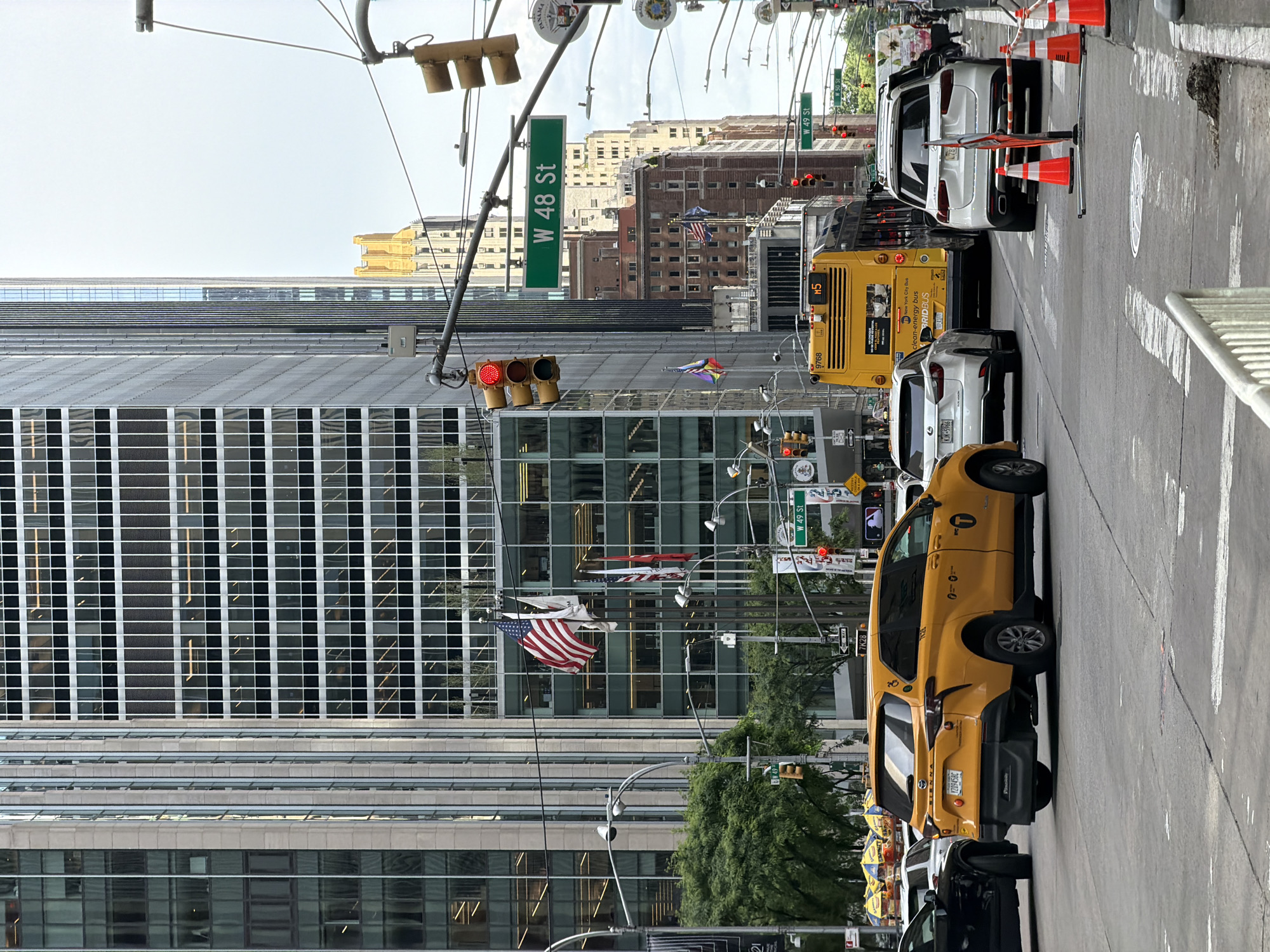 This image captures a vibrant street scene in a major city, likely New York City, oriented incorrectly by 90 degrees clockwise. The setting depicts a busy urban street lined with towering buildings and various vehicles under a bright, clear sky, suggesting daytime.
In the foreground, a yellow taxi, which appears to be a Toyota Sienna, is prominently parked on the right side of the street. Its license plate, "KJK-5966", is visible. Ahead of it, a white SUV is parked, followed by a yellow New York City bus. The bus displays "M5" as its route, "clean-energy bus", and "BRIDBUS" text, along with the number "9768". Further down the street, another white SUV and a dark-colored car are parked, with orange construction cones and barriers indicating ongoing work.
Tall, modern glass-fronted skyscrapers with intricate window patterns dominate the left side of the image, reflecting the surrounding environment. In the midground, an American flag and other flags are displayed from a building. Older brick buildings are visible further in the background.
Various street infrastructure elements are present, including traffic lights showing a red signal, utility poles with numerous wires, street lamps, and street signs. Notable street signs include "W 48 St" and multiple "W 49 St" signs. Other visible text includes "NAMA" on a small sign and possibly "7K28" on a vehicle or sign. The scene conveys a typical day in a bustling city environment. No specific event is unfolding, but rather the everyday activity of urban life.