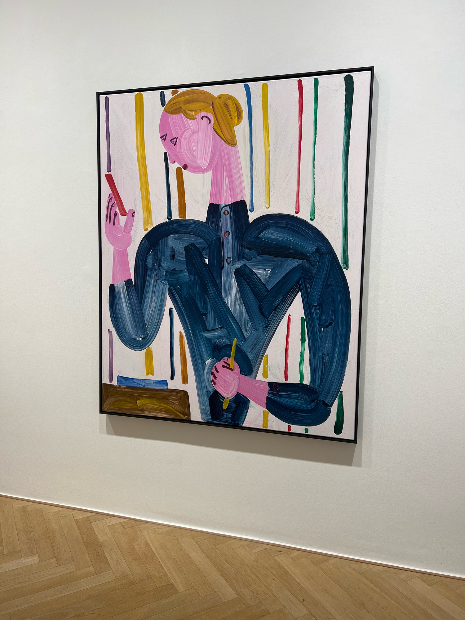 A framed, colorful painting hangs on a white wall in a gallery. The painting depicts a person with pink skin and blonde hair tied back in a bun, wearing a dark blue outfit. The person is holding two red sticks. The background of the painting is white with vertical lines of various colors, including yellow, red, green, purple, and brown. The floor is made of light brown wooden planks laid in a herringbone pattern. The image was taken in Berlin, Germany.