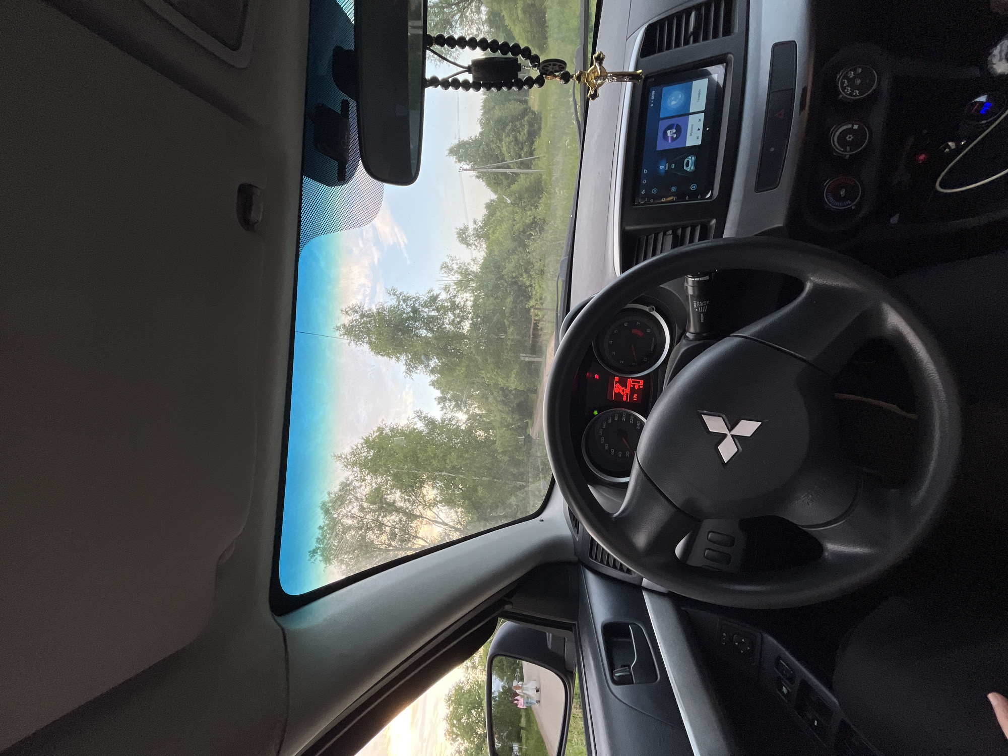 The image is a point-of-view shot from inside a car, looking through the windshield. The steering wheel and dashboard are prominent in the foreground, featuring a Mitsubishi logo on the steering wheel and a digital infotainment screen. Outside the car, a road winds through a tree-lined landscape under a cloudy sky with sunset hues. In the left side mirror, two figures are visible walking away on the road. The car appears to be stationary. The location is identified as Nikolskoye, Russia.