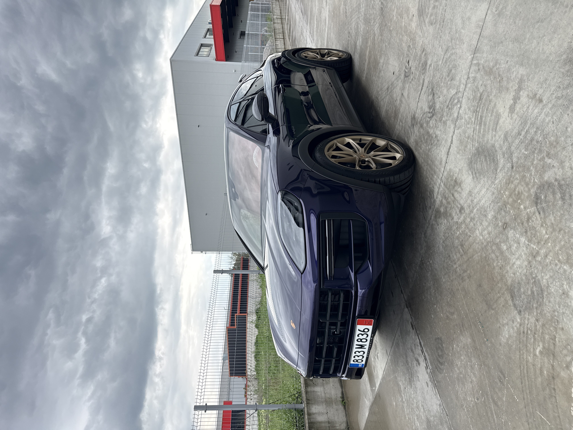 A dark blue Porsche Cayenne SUV is parked outdoors on a concrete surface under an overcast sky. The luxury vehicle is seen from a slightly low, front-side angle, facing left in the frame. It features distinctive gold-colored multi-spoke wheels fitted with Continental tires. The concrete ground exhibits subtle tire marks, suggesting recent movement.
In the background, a large, modern industrial or commercial building with light gray paneling and a striking red roof section stands under the cloudy sky. A chain-link fence runs parallel to the car, separating the concrete parking area from a strip of sparse grass and the building beyond. The sky is filled with dense, gray clouds, indicating a heavily overcast daytime setting with soft, diffused light.
The front of the SUV prominently displays a Bulgarian license plate, reading "BG 833 M 836". Small numeric details "08" and "23" are visible on the blue stripe of the plate, and "12" is on the red stripe. Partially visible text below the main number appears to say "OFFICIAL SALE" or similar. The scene is set in what appears to be a commercial or industrial zone, consistent with Plovdiv, Bulgaria.