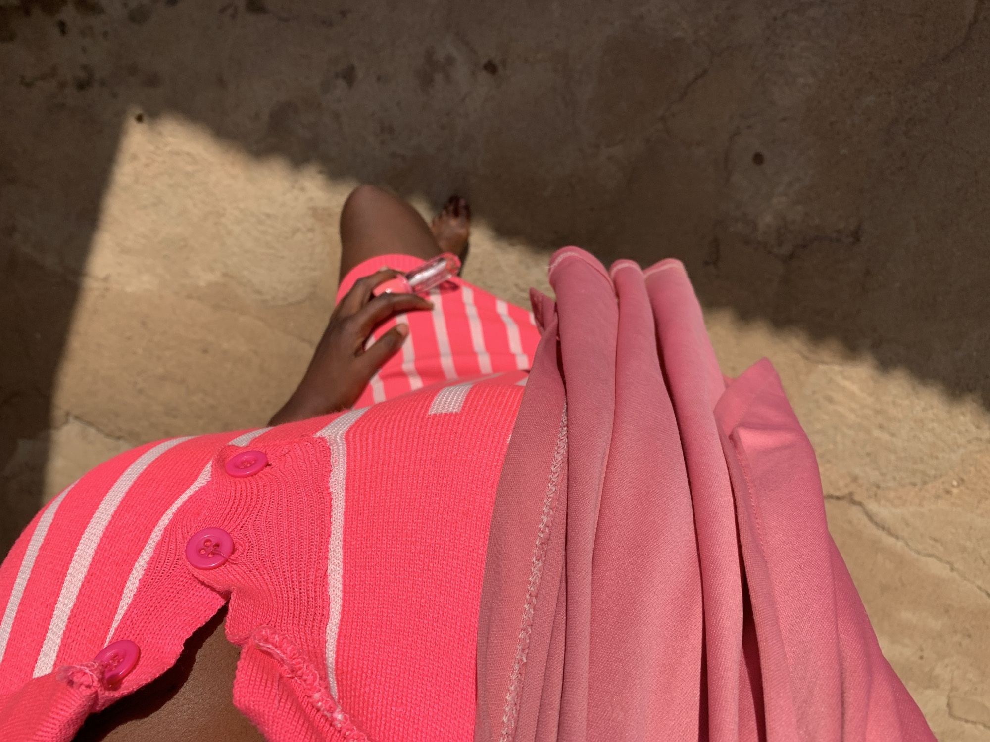 A downward-facing photograph shows a person seated outdoors in Rafin Par, Nigeria. The person is wearing a bright pink striped shirt and skirt, and holds a tube of clear lip gloss in their left hand. To the right, a soft, pink cloth is draped. The ground is light brown with darker brown shadows and cracks. A sliver of the person’s right foot is visible in the upper-left.