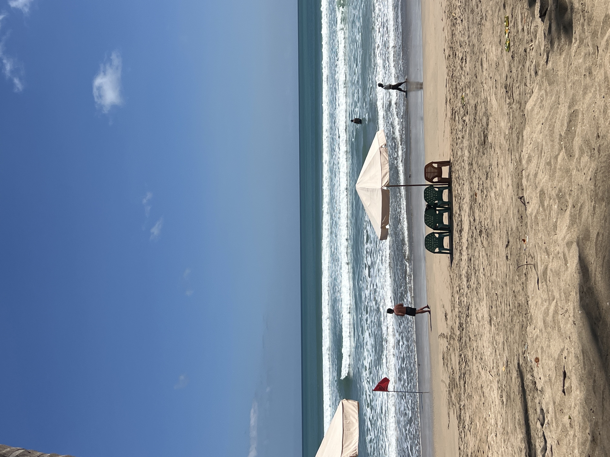 The image depicts a sunny day at Kuta Beach in Indonesia. The ocean has moderate waves and a clear blue sky with a few wispy clouds. On the sandy beach, there are several objects and individuals. Two large, light-colored beach umbrellas are visible, one on the left and another in the middle of the frame. In front of the middle umbrella, three beach chairs are set up: two green chairs and one brown chair. A red flag is planted on a pole near the water's edge, to the left of the central umbrella.
There are at least four people visible. A person wearing dark shorts is standing on the sand near the red flag, facing the water. Further out in the water, another person is standing or wading. On the right side of the frame, a person in a light-colored shirt and dark pants is walking along the shoreline. The sand itself has some debris scattered on it, including small leaves and twigs. The overall scene suggests a typical day at a popular beach resort.