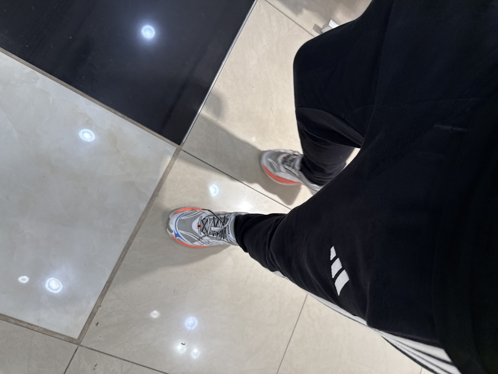 The image shows a first-person perspective of a person wearing black athletic pants with white stripes and grey and orange athletic shoes. The person is standing on a floor tiled with alternating light-colored and black tiles. Small circular lights are embedded in the floor tiles. The scene is indoors and appears to be a public space, possibly a shopping mall or a modern building lobby. The location is Stara Zagora, Bulgaria.