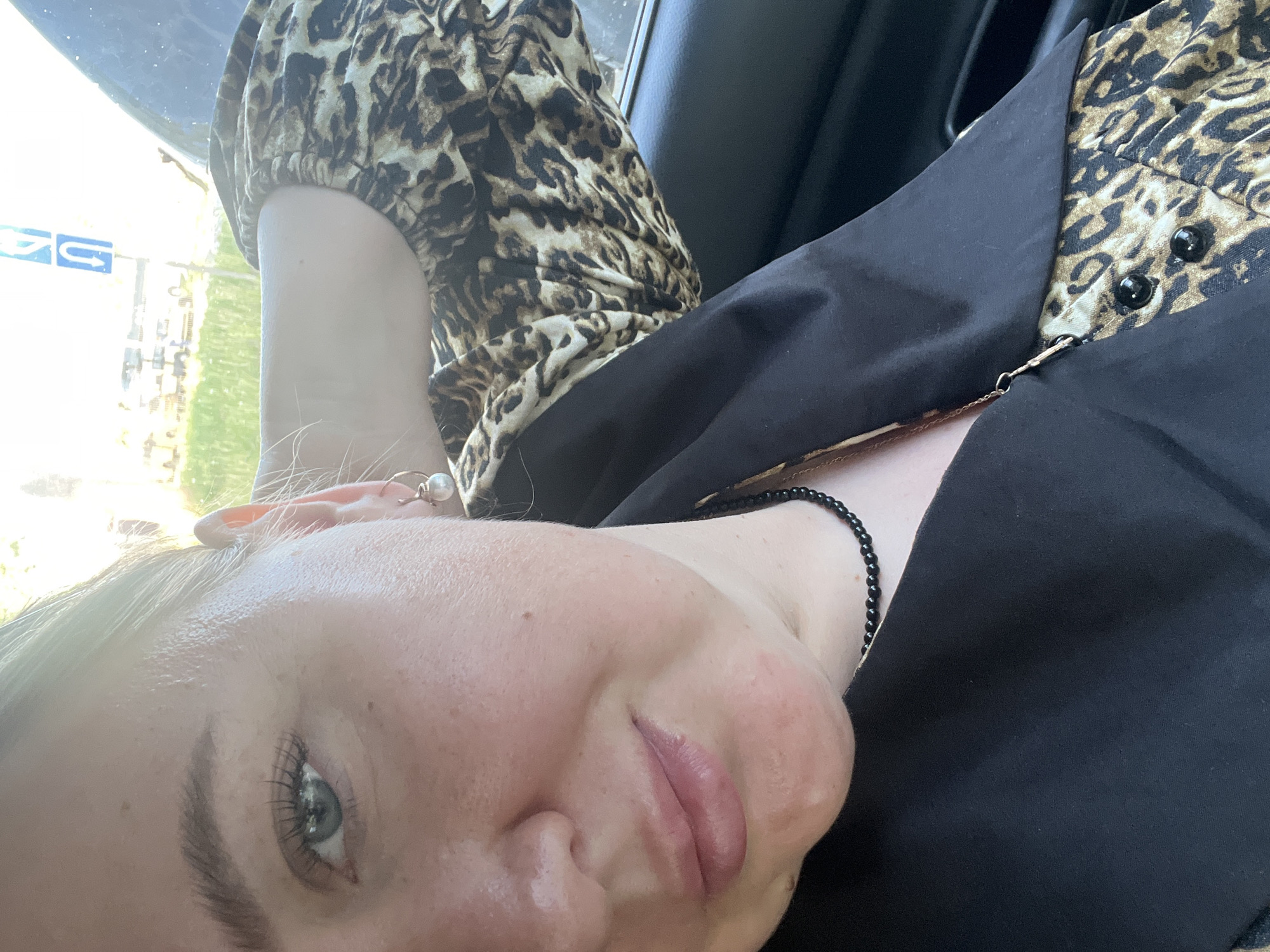 A selfie shot, presumably taken inside a car, shows a person with fair skin and blond hair. The person is wearing a leopard print top with puffed sleeves and a black blazer. A beaded necklace is visible around their neck, and they have a pearl earring. The background features the interior of the car, including a window and part of a car seat. Outside the window, a blurry sign with a blue emblem can be seen. While the location is given as Chaikovsky, Russia, there are no details in the image that explicitly confirm this location.